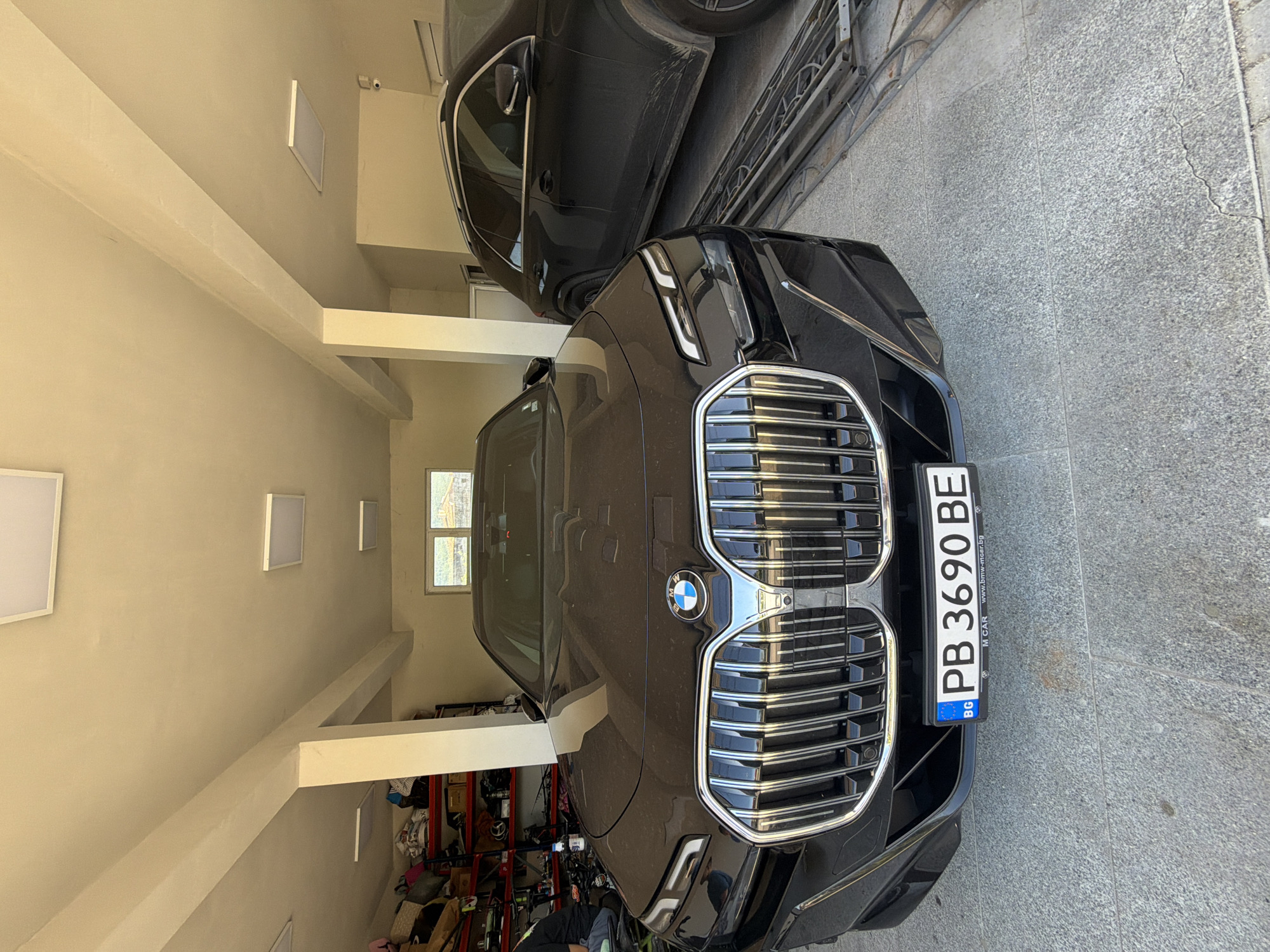 The image shows the front of a black BMW with a Bulgarian license plate "PB 3690 BE". The car is partially inside a garage or carport, with another dark-colored vehicle visible behind it. The garage interior includes supporting beams, and clutter on shelves in the background. The ground outside the garage appears to be paved. The location is plausibly Asenovgrad, Bulgaria, based on the license plate and the general architectural style. There are no people clearly visible in the frame, although a glimpse of a person wearing a dark jacket can be seen in the bottom of the image. The lighting is a mix of natural light coming from the front and artificial light within the garage.