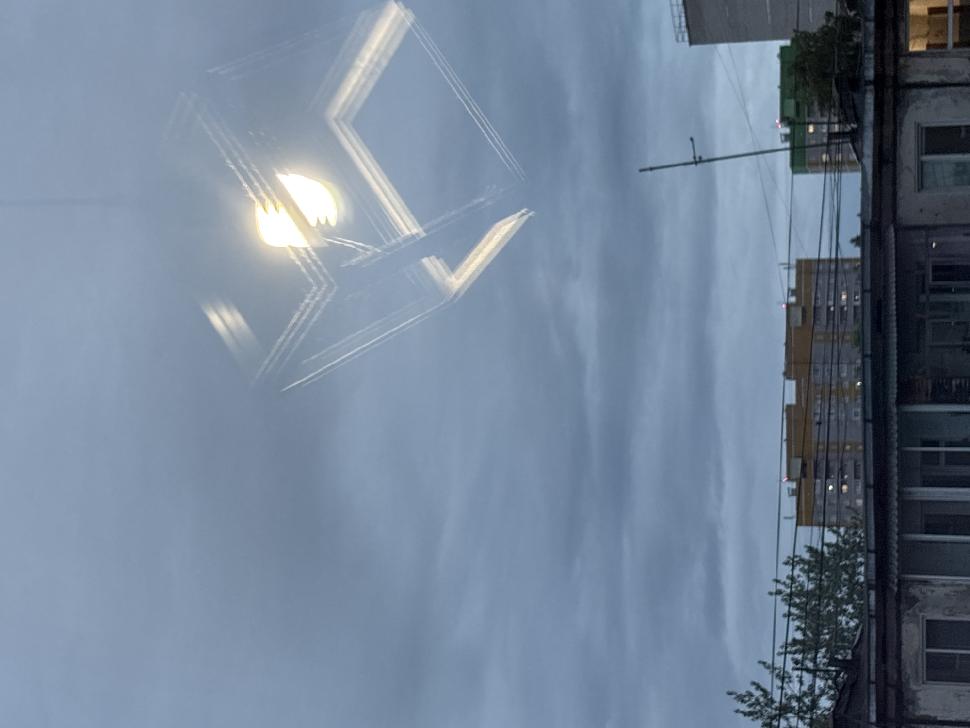 This photograph captures an urban landscape under an overcast sky, taken from an interior vantage point, exhibiting a prominent reflection of an indoor light source. The scene is located in the city of Izhevsk, Russia.
The upper portion of the image, approximately two-thirds, displays a uniformly grey, heavily clouded sky, indicating diffused ambient light. Superimposed on the upper right quadrant of this sky is a distinct reflection of an indoor light fixture. This fixture features a single, brightly glowing light bulb emitting a warm, yellowish light. The bulb is encased within an open, cuboid-shaped frame, rendered by bright, linear elements that create a three-dimensional effect. The reflection exhibits minor blurring and ghosting of these linear elements, characteristic of a surface reflection. A slender vertical element, likely a suspension wire or rod, extends upwards from the light source.
The lower third of the image reveals an urban environment. In the immediate foreground, the dark, corrugated roof and upper sections of a low-rise, possibly older building with multiple windows are visible. To the left, leafless tree branches partially obscure the view.
The midground is characterized by a dense network of dark utility lines stretching horizontally across the frame. Beyond these lines, several multi-story residential buildings are present. On the left, a structure with a light beige or yellow facade and a darker grey lower section features small red lights visible on its roofline. To the right, another residential building with a green upper section and a grey lower half also displays similar red roof-mounted lights. A tall, slender, dark mast or antenna ascends vertically from a rooftop on the right side of the midground. Pockets of green foliage from trees are scattered among these buildings.
No human figures or animals are observable, and no specific activities or interactions are depicted within the frame. The overall impression conveyed is one of a still, cloudy day in a residential district.
