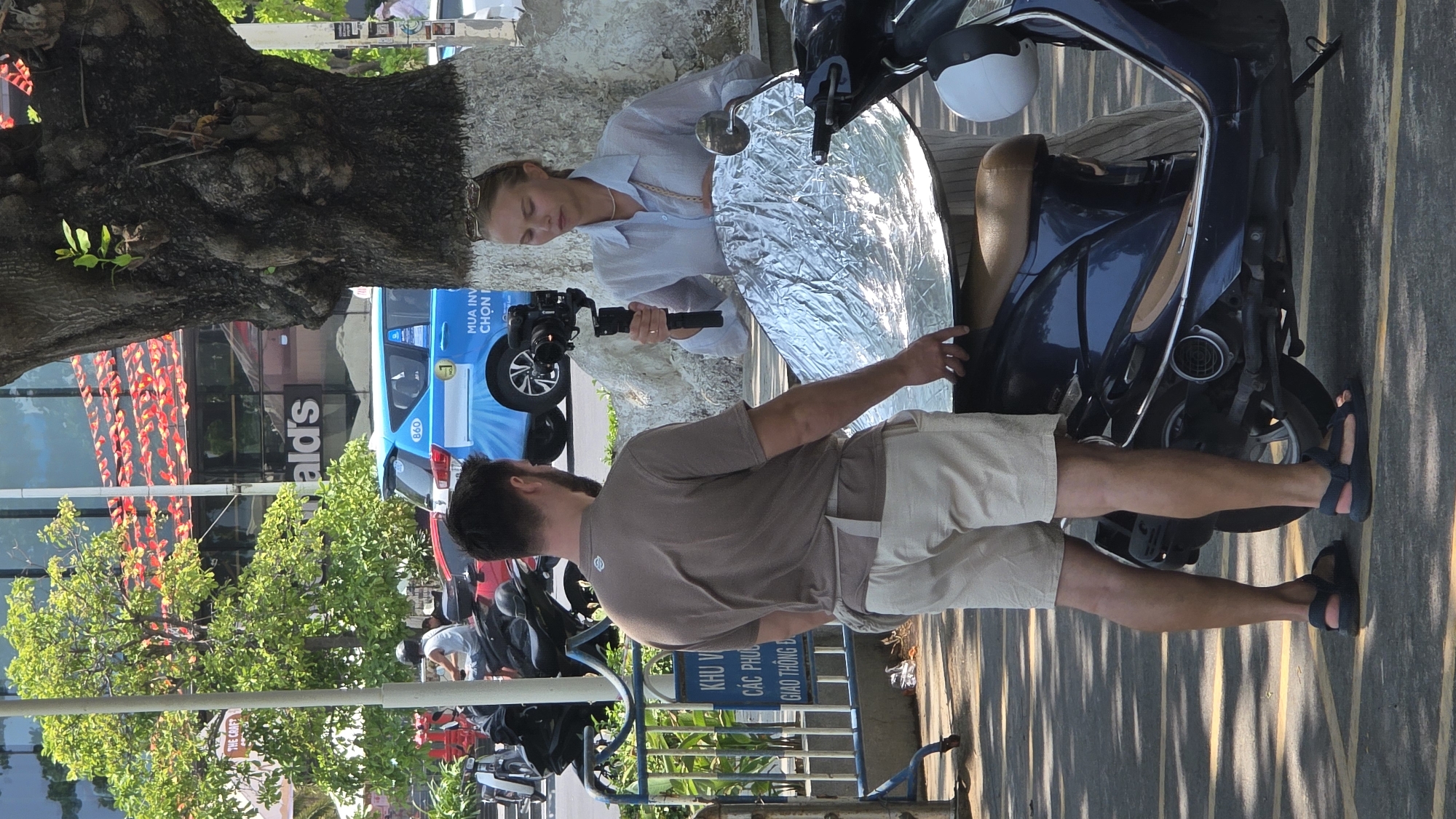 The image depicts a photoshoot occurring in Nha Trang, Vietnam, identifiable by the Vietnamese signage and recognizable features in the background. A fair-skinned female model with blonde hair is positioned near a parked blue motorbike and a reflector, with a camera held in her hands. An individual in a brown t-shirt and beige shorts stands behind the model. The scene is outdoors, with a large tree visible on the left side of the frame. In the background, a McDonald's restaurant and various vehicles, including a blue and white van, are visible. The street has painted yellow lines.