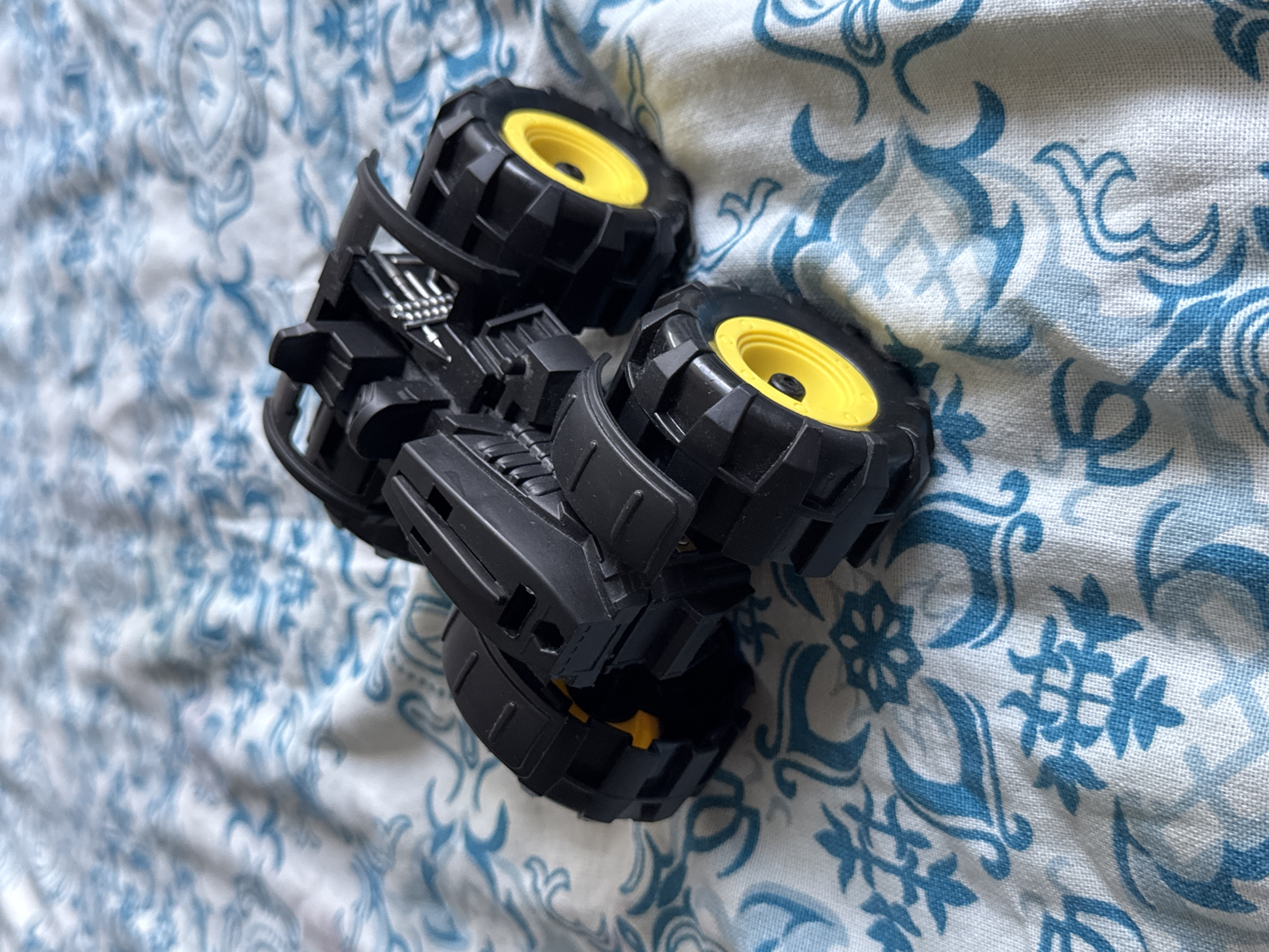 A black toy monster truck with yellow wheels is positioned on a white fabric surface patterned with blue floral designs. The truck is viewed from a slightly elevated, angled perspective, with its front to the right and slightly elevated. The toy is in focus, while the background is slightly blurred. The scene appears to be indoors, on a bed or similar soft surface, in the Яромінскі сельскі Савет, Belarus.