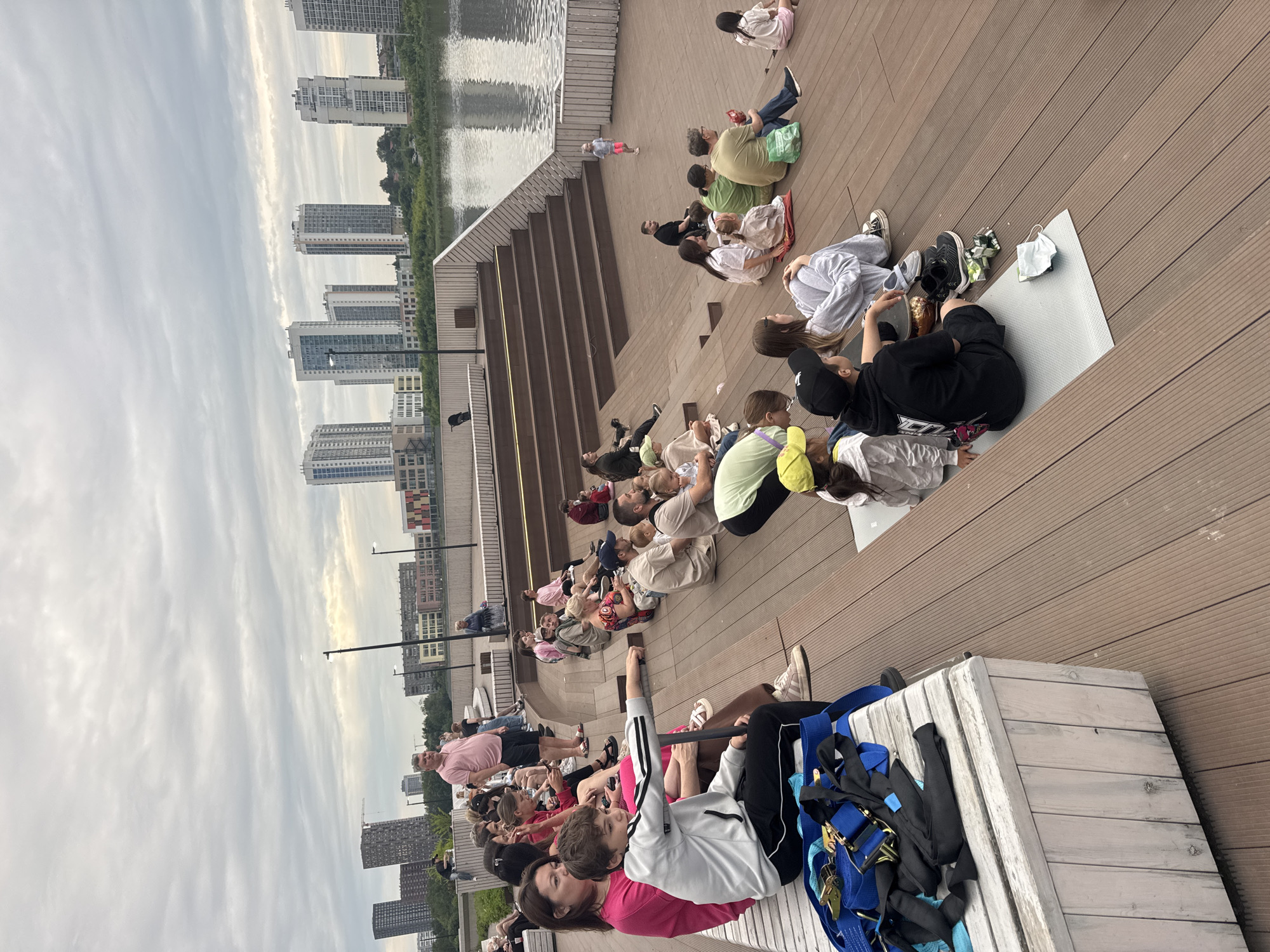 An outdoor amphitheater with tiered wooden seating is filled with people watching an event. The seating area is constructed from dark brown wooden planks and overlooks a body of water, likely a river or lake, with a city skyline of tall buildings in the background. The sky is overcast with scattered clouds.
In the foreground, a group of people are seated on the wooden steps. To the left, a white wooden bench is partially visible, with a pile of blue and black straps on it. A young boy in a white hoodie and black pants is seated on the steps, leaning his arm on a railing. Several other individuals, including women and children, are seated around him.
Further up the amphitheater, more people are visible, scattered across the seating area. Some are standing and looking towards the front of the amphitheater, while others are seated.
In the middle ground, a person in a pink shirt is standing and appears to be speaking or directing.
In the background, across the water, a modern city skyline is prominent, featuring numerous high-rise buildings. The architecture suggests a contemporary urban setting. The overall scene depicts a public gathering in an urban park or recreational area, possibly a performance or lecture, taking place on a cloudy day. The presence of modern high-rise buildings in the background suggests this location is within a city. Given the style of architecture and the inclusion of a large body of water, this scene could be Yekaterinburg, Russia, known for its urban development and proximity to water bodies.