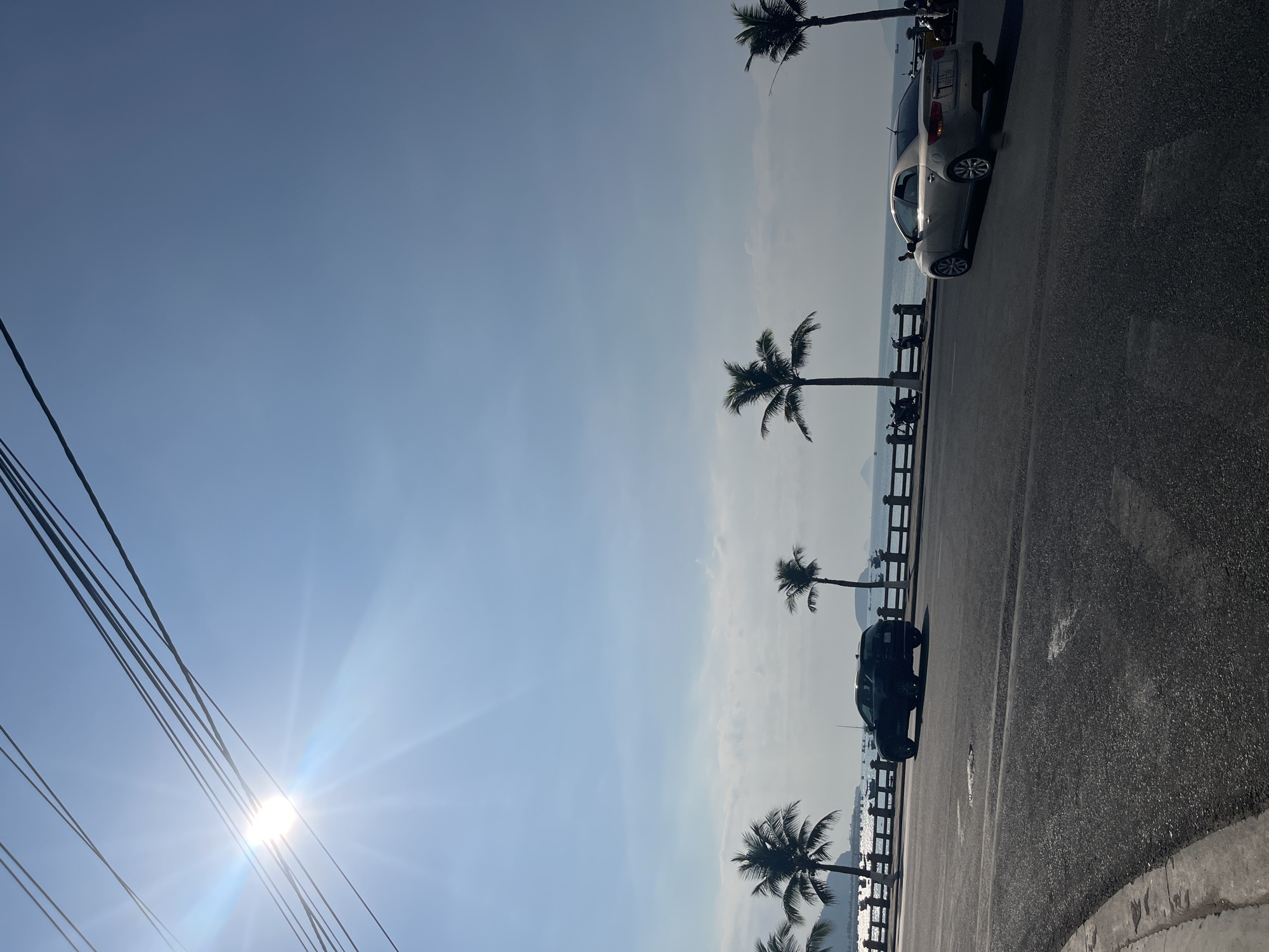 The image depicts a coastal scene in Bac Nha Trang Ward, Vietnam. A sunny sky is visible with a bright sun partially obscured by electrical wires. The ground area includes a paved road and a sidewalk with a dark-colored vehicle and a silver car parked. The background shows a view of the sea with a wooden fence separating the road from the sea view. Several palm trees are visible along the coast. People are seen standing near the cars.