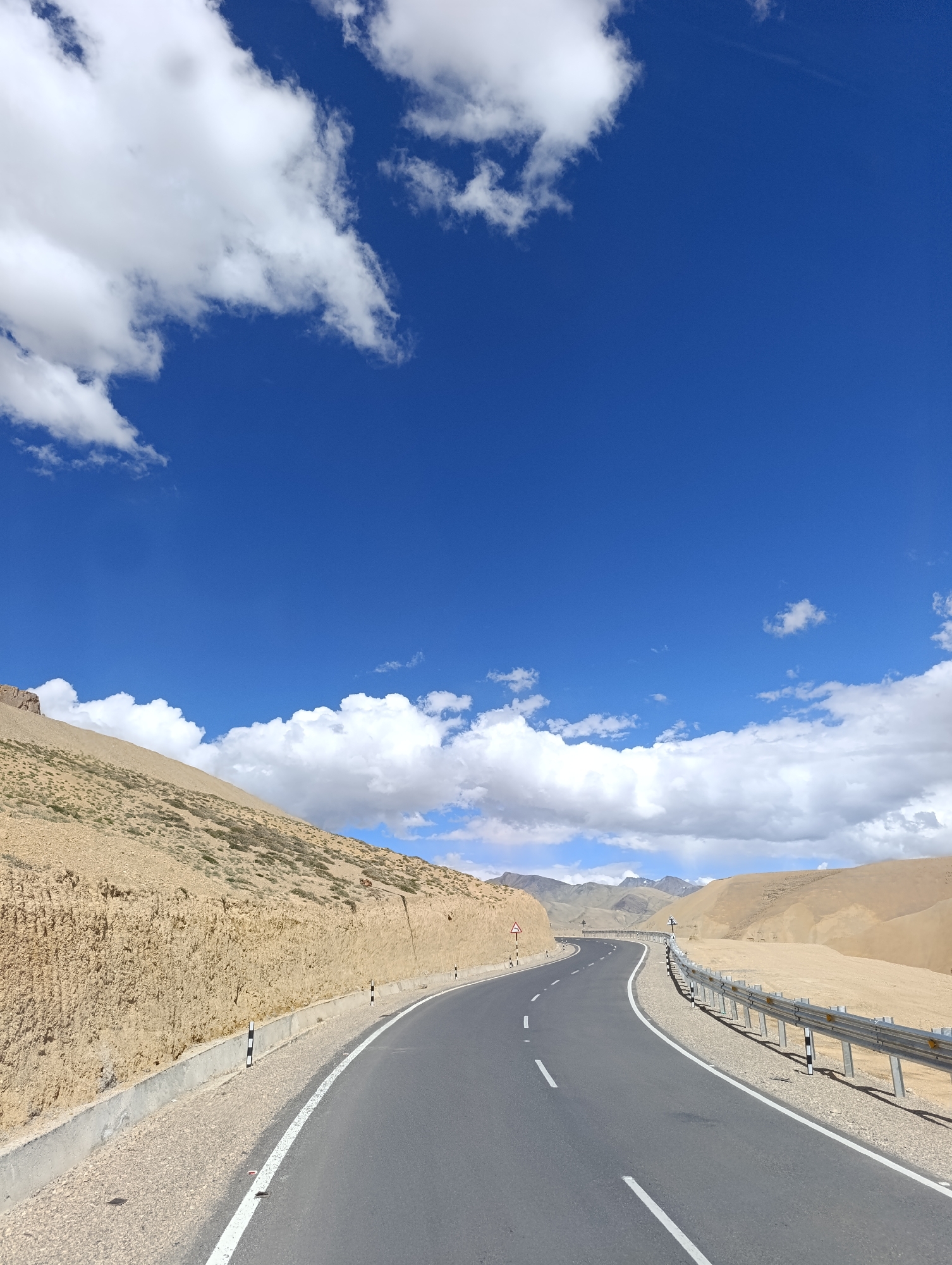 A scenic view of a winding asphalt road cuts through a mountainous, arid landscape under a vast blue sky dotted with fluffy white clouds. The road, marked with solid white lines on the right and broken white lines on the left, curves to the right, disappearing into the distance. On the left side of the road, a steep, sandy embankment is carved out, revealing layers of earth. Along the right edge of the road, a metal guardrail with silver posts and reflective markers runs parallel to the tarmac. Beyond the guardrail, the terrain is a gently sloping, barren expanse of sand and rock. In the far distance, a range of majestic, rugged mountains with muted brown and grey tones forms the horizon. The overall scene is one of natural beauty and a remote, high-altitude environment.