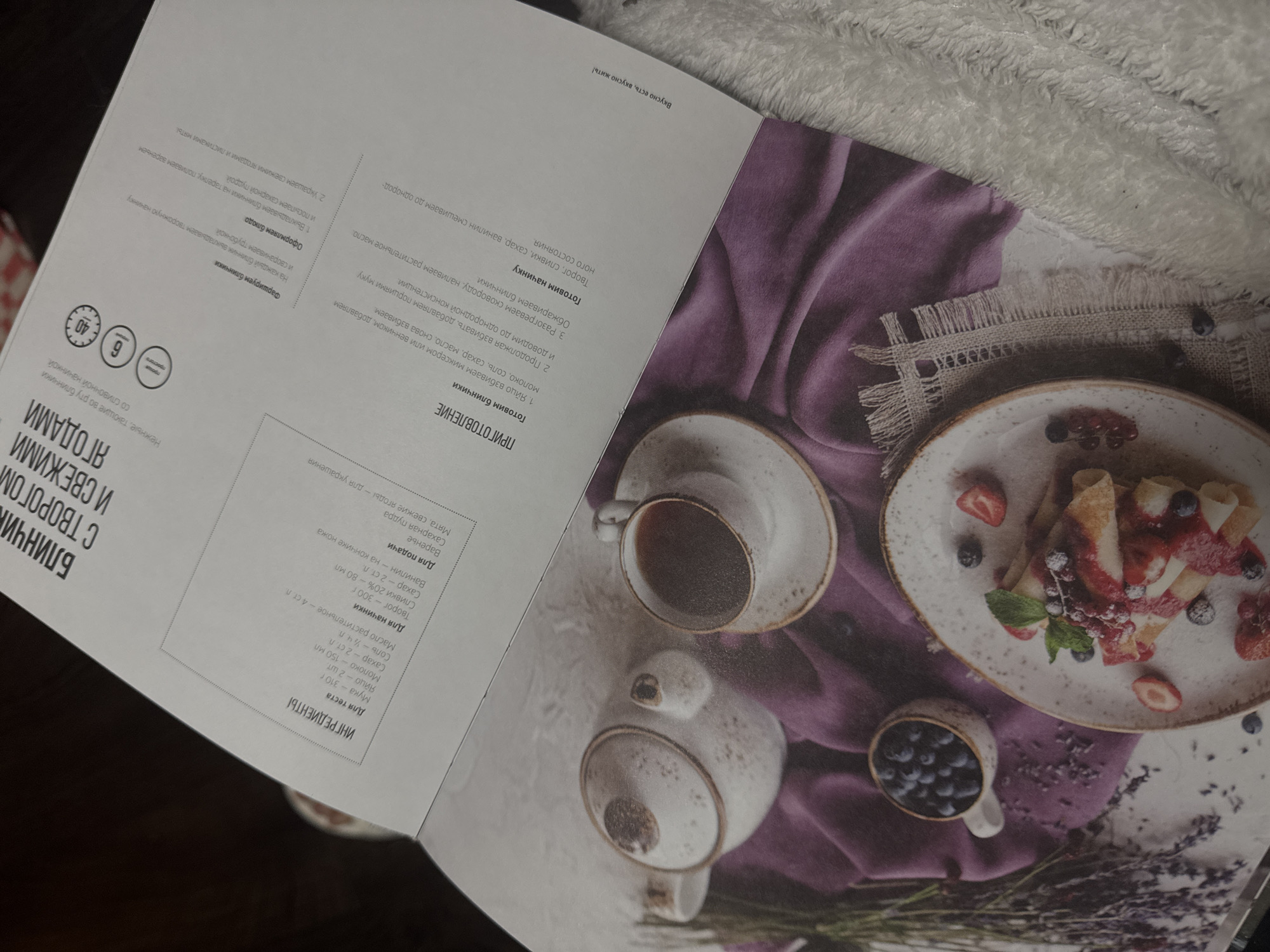 The image shows a recipe spread from a cookbook or magazine. The left side features a list of ingredients and instructions for making crepes with cottage cheese and fresh berries. The right side has a stylized food photography scene with a rustic aesthetic. There is a white plate with rolled crepes topped with strawberry jam, fresh strawberries, blueberries, and mint leaves. A cup of coffee and a teapot are also present. Lavender sprigs are placed around the food. The scene is set on a dark surface with a violet-colored tablecloth underneath the food.