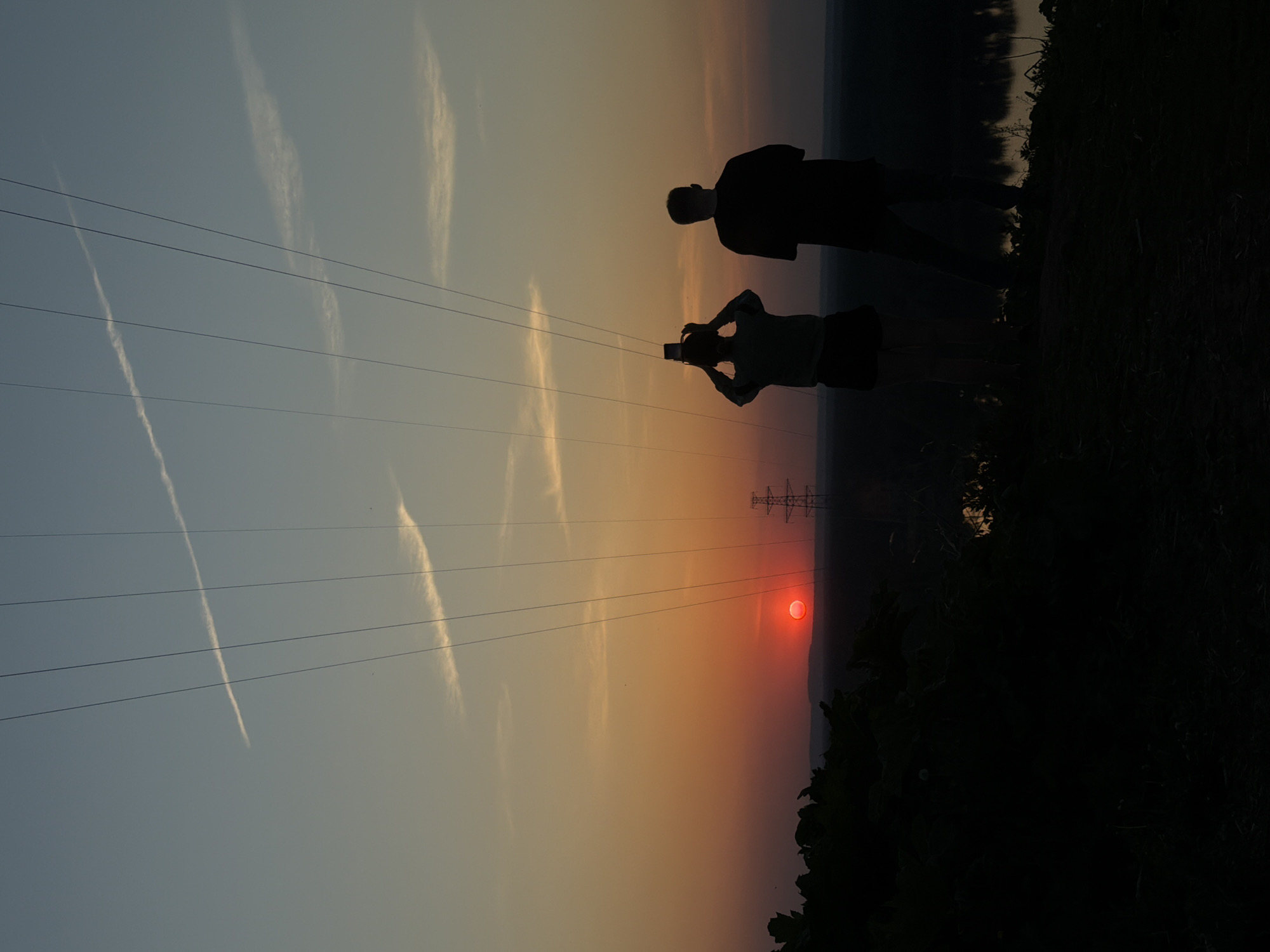 The image depicts a sunset scene in Noviy, Russia. Two silhouetted figures stand on what appears to be a grassy embankment. One figure holds a mobile phone up, seemingly taking a photograph. An electrical tower is visible in the background, positioned near the setting sun, which appears as a red orb. Overhead power lines are strung across the view. The sky is a gradient of colors, from a pale blue to an orange-red near the horizon. Contrails from an aircraft are visible in the sky. The foreground vegetation is dark, bordering the right edge of the image.