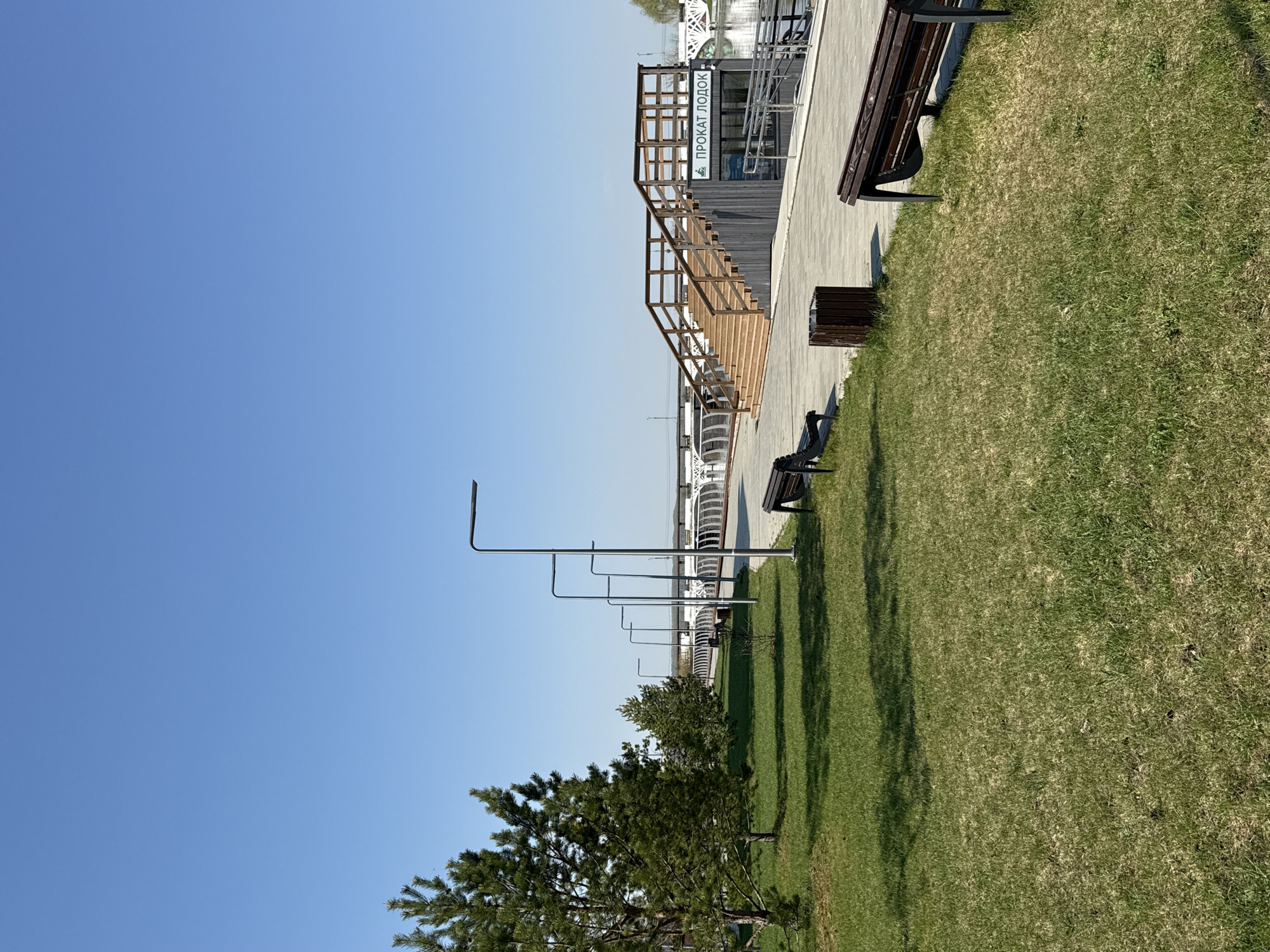 A low-angle, medium shot shows a promenade in Votkinsk, Russia on a clear, sunny day. In the top right corner, a wooden bench sits on grass. Moving left, a wooden trash receptacle stands adjacent to a walkway. A row of several metal benches extends towards the middle of the frame. Next to the walkway, a wooden building with stairs leading up to a second level. A sign reading "ПРОКАТ ЛОДОК" (boat rental) in Russian is visible on the building. Further in the background, white cylindrical structures line the walkway. Along the bottom, green grass and trees are visible. Street lamps punctuate the walkway. The sky is clear and blue.