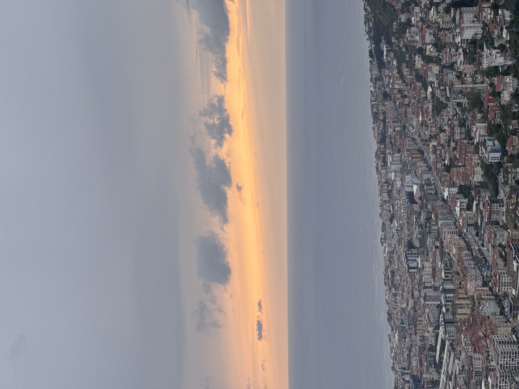 The image depicts a cityscape next to the ocean under a sky with varying cloud cover and a golden hue, possibly during sunset or sunrise. The city is densely populated with numerous buildings, some of which are adjacent to trees. The ocean's surface appears calm.