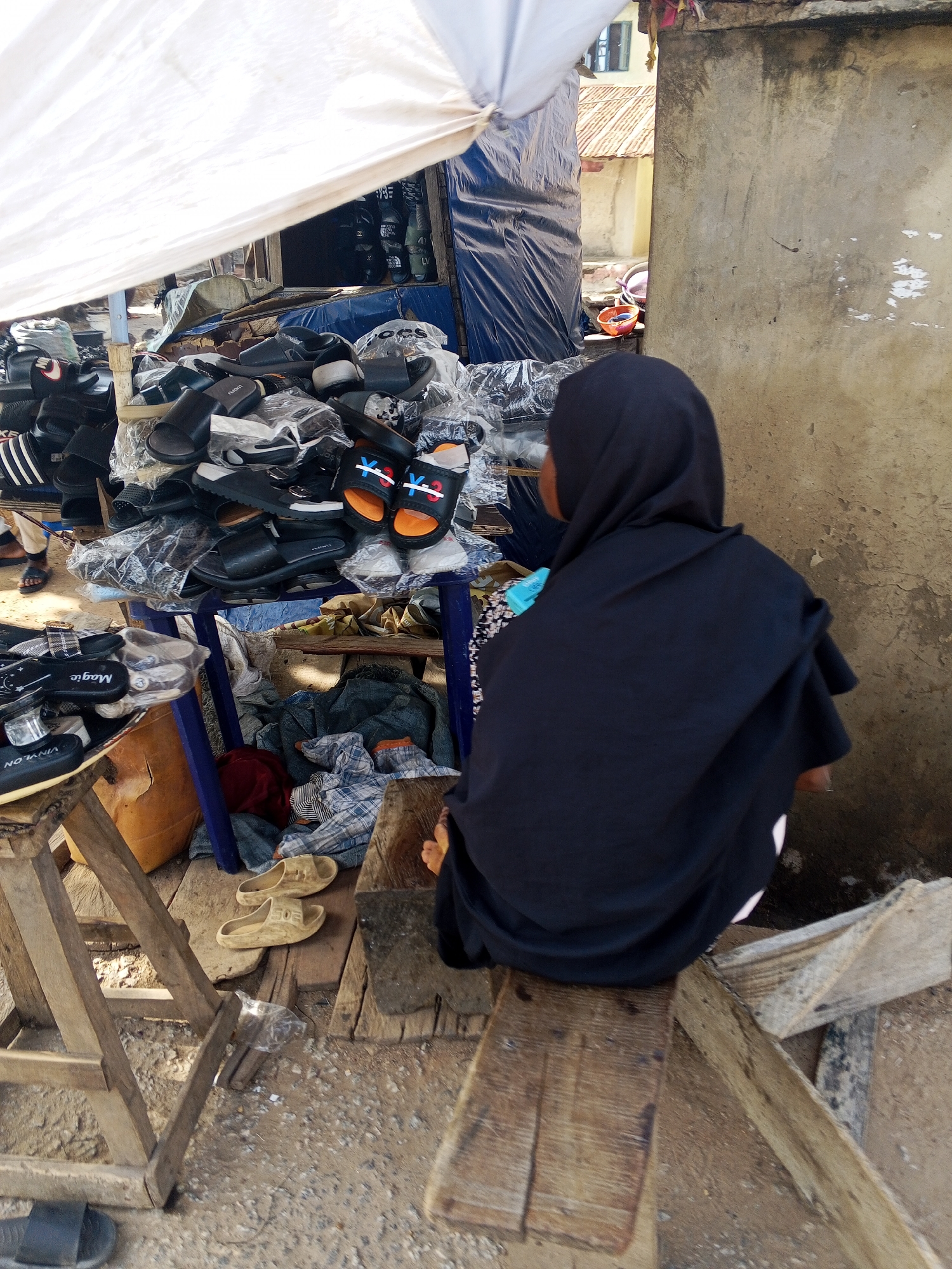 The image depicts an outdoor market scene in Adankolo, Nigeria, featuring a vendor and a shoe stall under a white tarp. A person, seen from behind, wears a dark head covering and dark garment, seated on a simple wooden bench. They appear to be attending the stall, which is piled high with various sandals and slippers.
The footwear is displayed on a blue plastic table and other wooden structures, with many pairs still in clear plastic packaging. Visible brands and text on the sandals include "Magic," "VINYLON," and the three stripes logo of "Adidas." Some sandals feature distinctive "X" and "Y" or "XX" and "YY" designs, while others in the background have logos resembling "LV." Underneath the main display, there are folded clothes (including a plaid pattern), a reddish fabric, and a brown container.
The ground is unpaved, a mix of dirt and gravel, with scattered debris. In the background, a weathered concrete wall and glimpses of buildings with corrugated iron roofs provide contextual cues for the market setting. The bright, clear daylight suggests it is daytime, with strong shadows visible.