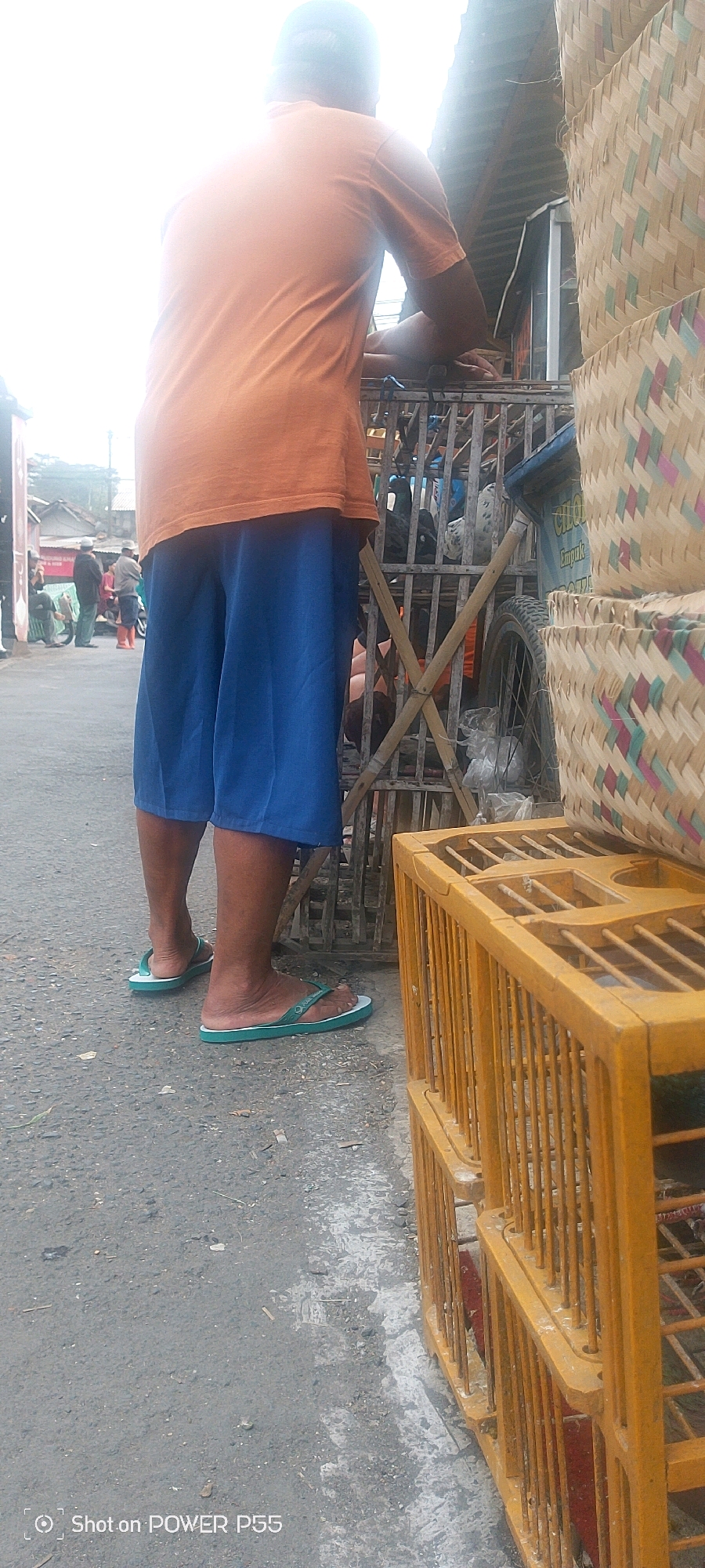 In an outdoor, urban setting, a person stands facing away from the camera, wearing an orange t-shirt and blue shorts. They are barefoot in green flip-flops. The person appears to be leaning on a wooden structure, possibly a fence or stall. To the right of the person, there are several stacked woven baskets made of bamboo and colored with pink, green, and blue accents. Below the baskets, two yellow bird cages are stacked, constructed from wooden bars. One of the cages contains what appear to be pigeons. Behind the person, a dirt or asphalt road is visible, with a few other people and buildings in the background. The lighting suggests it is daytime, with a bright sky. The ground is a mix of asphalt and some scattered debris. A water mark at the bottom of the image reads "Shot on POWER P55".