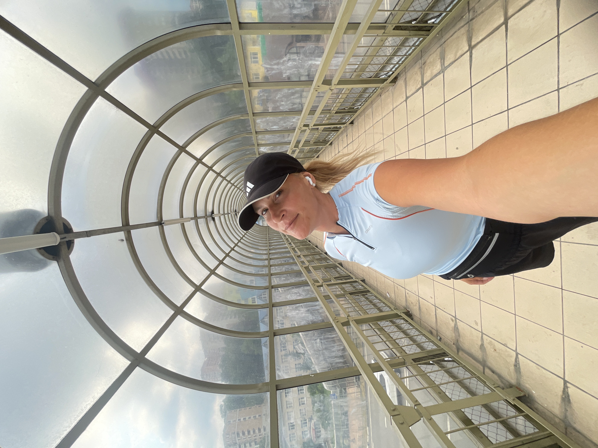 A woman is taking a selfie in a covered pedestrian walkway in Sochi, Russia. She is wearing a light blue athletic shirt, black shorts, and a black baseball cap. She has blonde hair and is wearing white wireless earbuds. The walkway has a curved, transparent roof supported by metal beams and a metal railing runs along the side. The floor is tiled. Through the transparent roof and sides, buildings and greenery are visible, suggesting an urban or suburban setting. The sky appears overcast. The lighting indicates it is daytime. The woman appears to be smiling and is looking at the camera. The overall mood seems casual and active, possibly indicating she is out for a run or a walk. The text "CRAFT" is visible on her shirt.