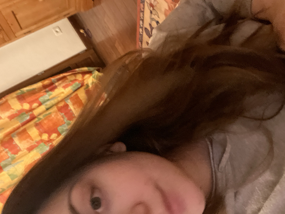 A close-up, tilted shot focuses on a person with long, brown hair partially covering their face. One visible eye is brown. The individual appears to be lying down. A grey garment is visible at the neck. In the background, there's a glimpse of a wooden cabinet and a bedspread with a pattern of orange, yellow, and green. The perspective is off-kilter, giving a disoriented feeling.