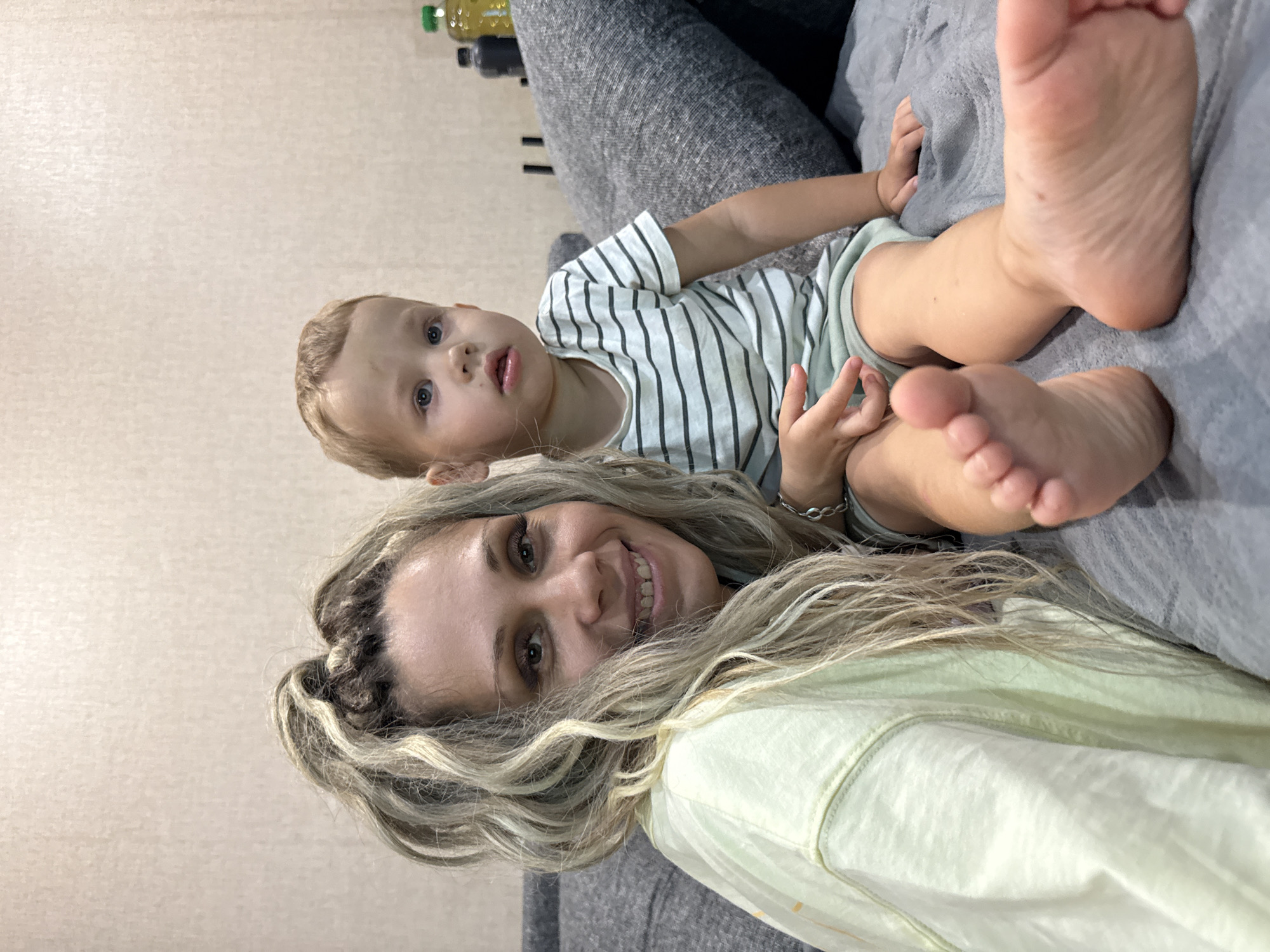 A woman and a young child are sitting on a couch. The woman, on the left, has long blonde hair in braids and is wearing a light yellow t-shirt. She is smiling at the camera. The child, on the right, has short blonde hair and is wearing a striped t-shirt and light shorts. The child is looking slightly off-camera. Their feet are visible in the foreground, with the child's bare feet prominently displayed. The background appears to be an indoor setting with a textured wall. There are some objects on a surface behind the couch, including bottles. The couch is upholstered in grey fabric. There are no discernible elements indicating the specific location of Rusanovo, Russia within the image.