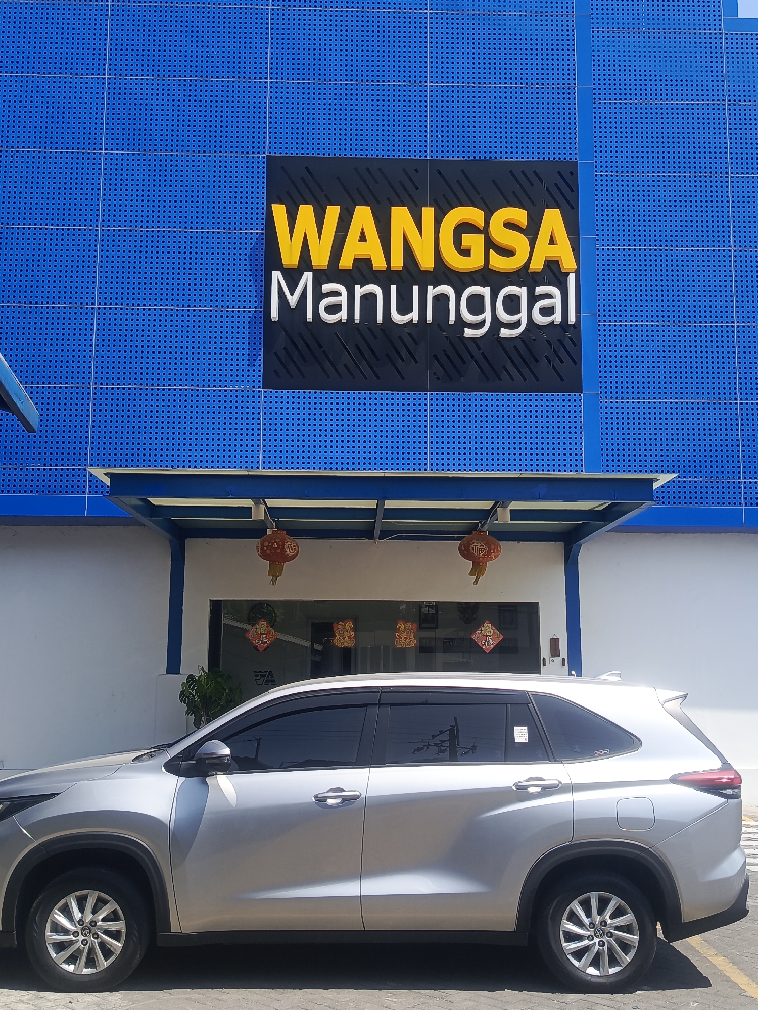 The image shows a silver SUV parked in front of a building with a blue facade. The building has a sign with yellow and white lettering that reads "WANGSA Manunggal". There are red Chinese lanterns hanging from the awning above the entrance. The car is viewed from the side, showing its front and rear wheels, doors, and tinted windows. The background consists of the building and a white wall. The scene appears to be outdoors, likely in a commercial area. The location is Wadungasih, Indonesia.