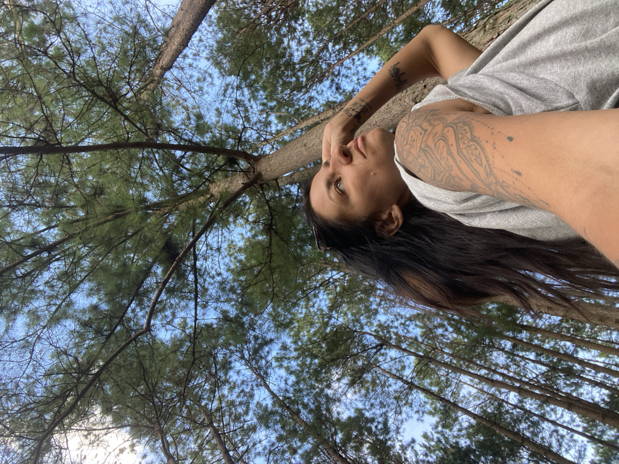 A woman is lying down, looking up at the sky through tall trees. The scene is outdoors, with a bright blue sky visible through the branches and pine needles of the trees. The woman has long, dark hair and is wearing a grey t-shirt. Her arm is resting on her head, and she has tattoos on her arm and shoulder. The image is taken from a low angle, looking upwards. It appears to be daytime, and the weather is clear. The overall mood is relaxed and contemplative. There are no visible text elements in the image.