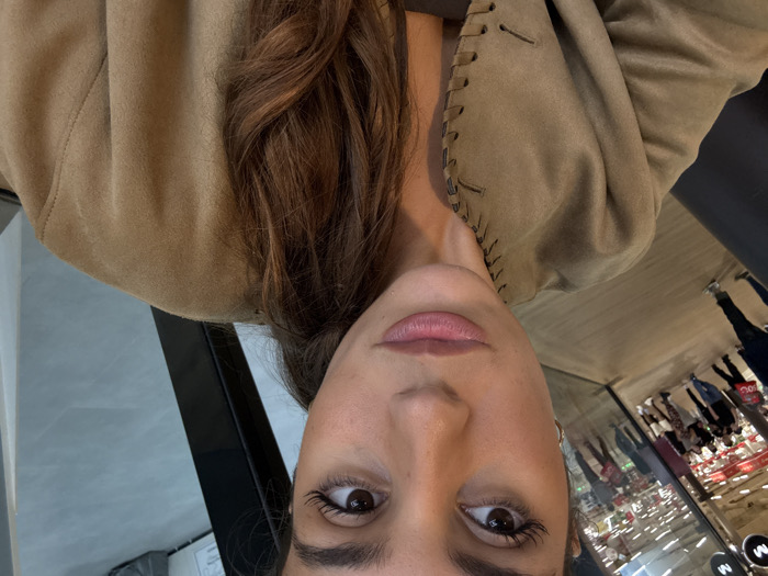An adult female is depicted from the chest up, occupying the right half of the frame, looking directly forward with a neutral expression. Her features include brown eyes with visible eyelashes, defined eyebrows, and light-toned lips. Her long, wavy brown hair rests primarily over her right shoulder. She is dressed in a light brown or tan jacket with distinctive lacing detail along the front opening. A darker garment is visible beneath.
The background consists of a large reflective glass surface. The left side of the reflection shows a busy retail environment with multiple individuals walking within a store aisle. Shelves are visible, stocked with products featuring red and white branding. A faint circular "M" logo is discernible in the upper left of this reflection, and the reflected floor appears light-colored. The far right background features a dark vertical structural element adjacent to a light grey wall or floor surface. This scene is located within a commercial establishment in Aldaia, Spain.