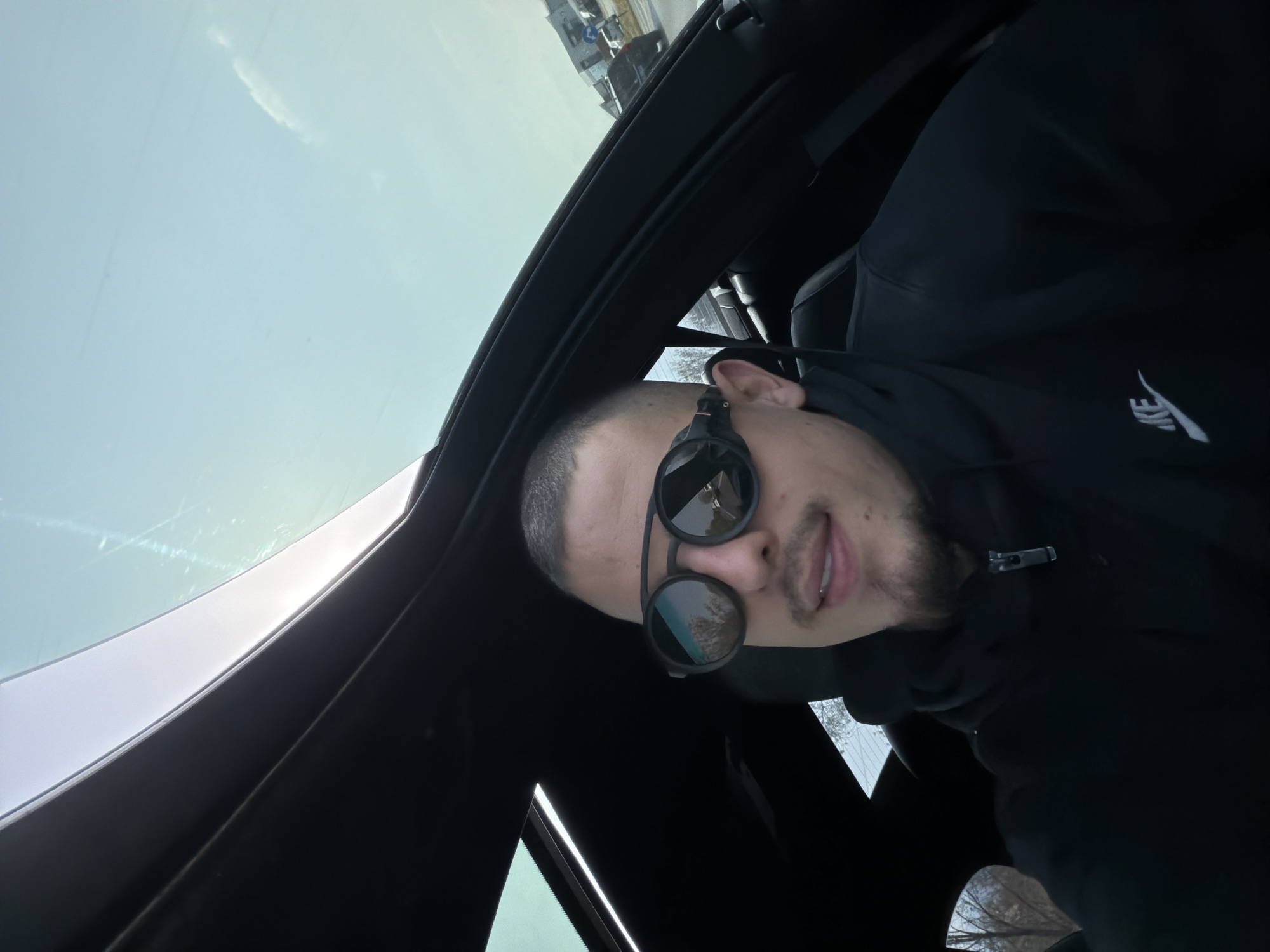 A man is photographed inside a car. He is wearing black sunglasses and a black hooded sweatshirt with a Nike logo on the chest. The car's sunroof is open, revealing a bright sky and what appears to be a street scene through the windshield. The reflection in his sunglasses shows trees. The location cannot be determined to be Plovdiv, Bulgaria from the visual information provided.
