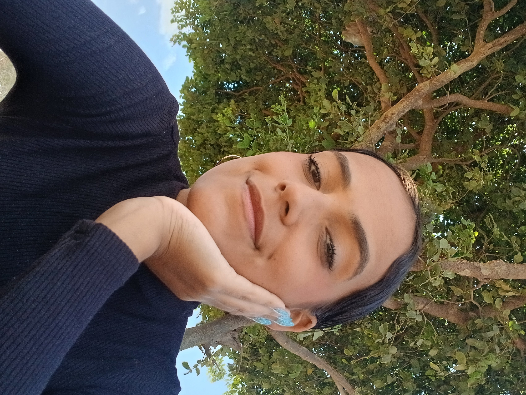 A close-up, rotated portrait captures a young woman outdoors, seemingly taking a selfie while resting her chin on her right hand. She has a relaxed expression with a gentle smile and eyes gazing towards the camera. Her dark hair is styled away from her face, revealing gold hoop earrings. She wears a dark, ribbed, long-sleeved top, and her fingernails are painted a sparkly light blue.
The background is dominated by a lush green tree canopy with visible branches, creating a natural and verdant setting. A patch of clear blue sky with some white clouds can be seen towards the upper left, indicating a bright daytime setting. The natural light illuminates her face and the foliage. The overall atmosphere is peaceful and serene. The image's orientation is ninety degrees clockwise. This scene is located in Altagracia, Venezuela. No visible text is present.