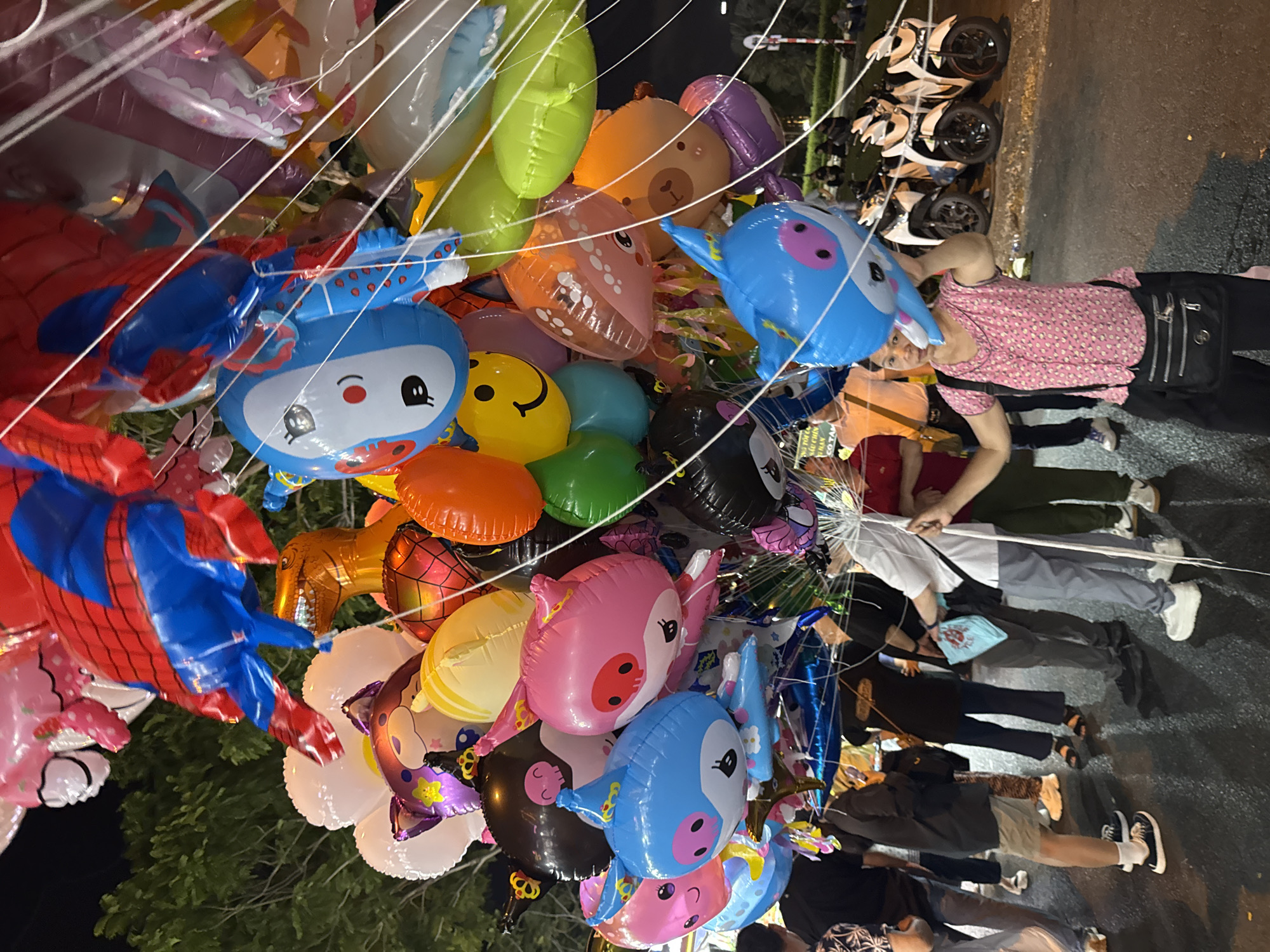 The image shows a close-up of a large bunch of balloons, predominantly character-themed (such as Spiderman, dinosaurs, and cartoon faces). The balloons are of various colors and shapes. In the background, several people are visible, standing on what appears to be a paved street. In the distance, some motorbikes are parked. It is nighttime. The scene appears to be taking place in a busy area, likely a street market or fair. The general environment suggests the location is in Asia; the city is identified as Đà Nẵng, Vietnam. There's a vendor holding the balloons, likely trying to sell them. The other people are presumably potential customers or passersby. The specific interactions are difficult to discern in detail.