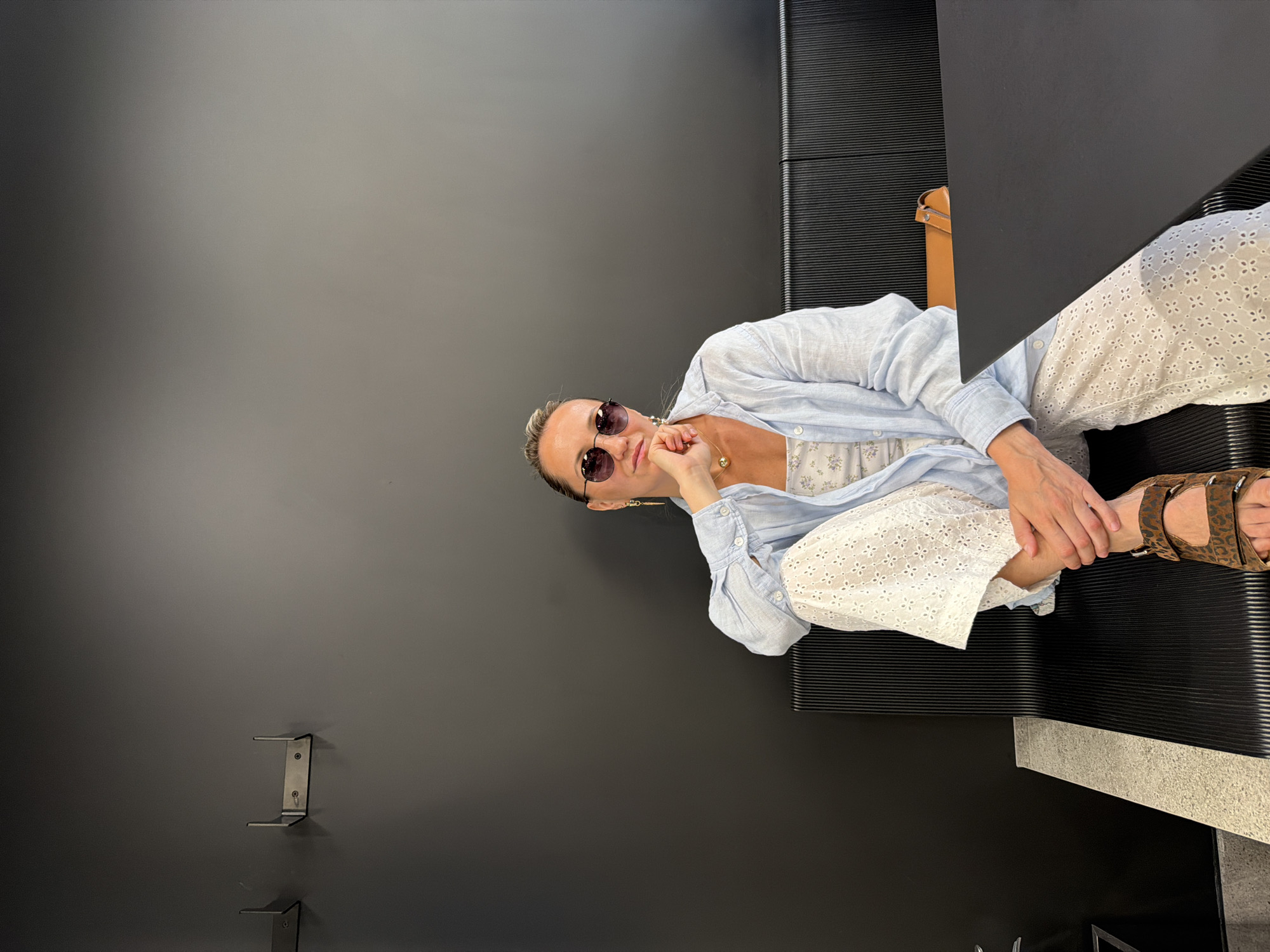 A woman with blonde hair pulled back sits on a black slatted bench against a dark gray wall. She is wearing a light blue unbuttoned shirt over a white floral camisole, white eyelet pants, and leopard print sandals. She has on round sunglasses and gold earrings. Her right hand rests on her chin. A brown handbag is visible on the right side of the bench. The scene appears to be indoors, possibly a cafe or a waiting area, with minimal natural light. There are two metal brackets mounted on the wall above and to the left of the woman. The overall aesthetic is modern and minimalist. The location of this scene in Izhevsk, Russia, is not determinable from the image alone.