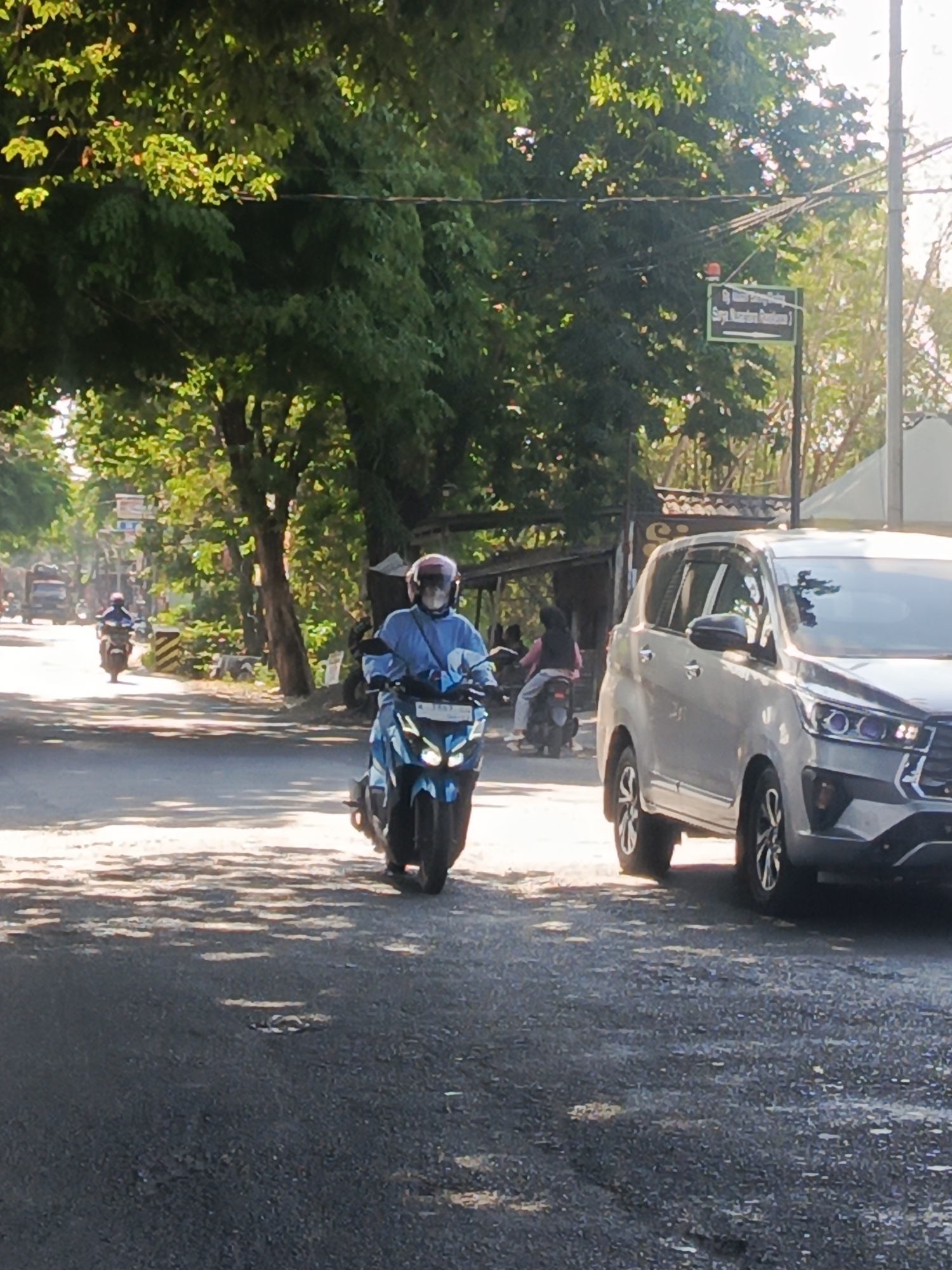 The image captures a street scene in Sidowungu, Indonesia, bathed in dappled sunlight filtering through lush trees lining the road. A person wearing a blue shirt and a helmet rides a blue scooter forward, their face obscured by the helmet's visor. To the right, the front quarter of a silver SUV is visible, parked or moving on the road. In the background, other vehicles, including motorcycles and a truck, are seen on the road, suggesting moderate traffic. A signpost with green text on a dark background is affixed to a pole amidst the foliage on the right side of the road. The asphalt surface of the road shows signs of wear, with several potholes visible in the foreground. The overall atmosphere is that of a typical day on a tree-lined street in a semi-urban Indonesian setting.