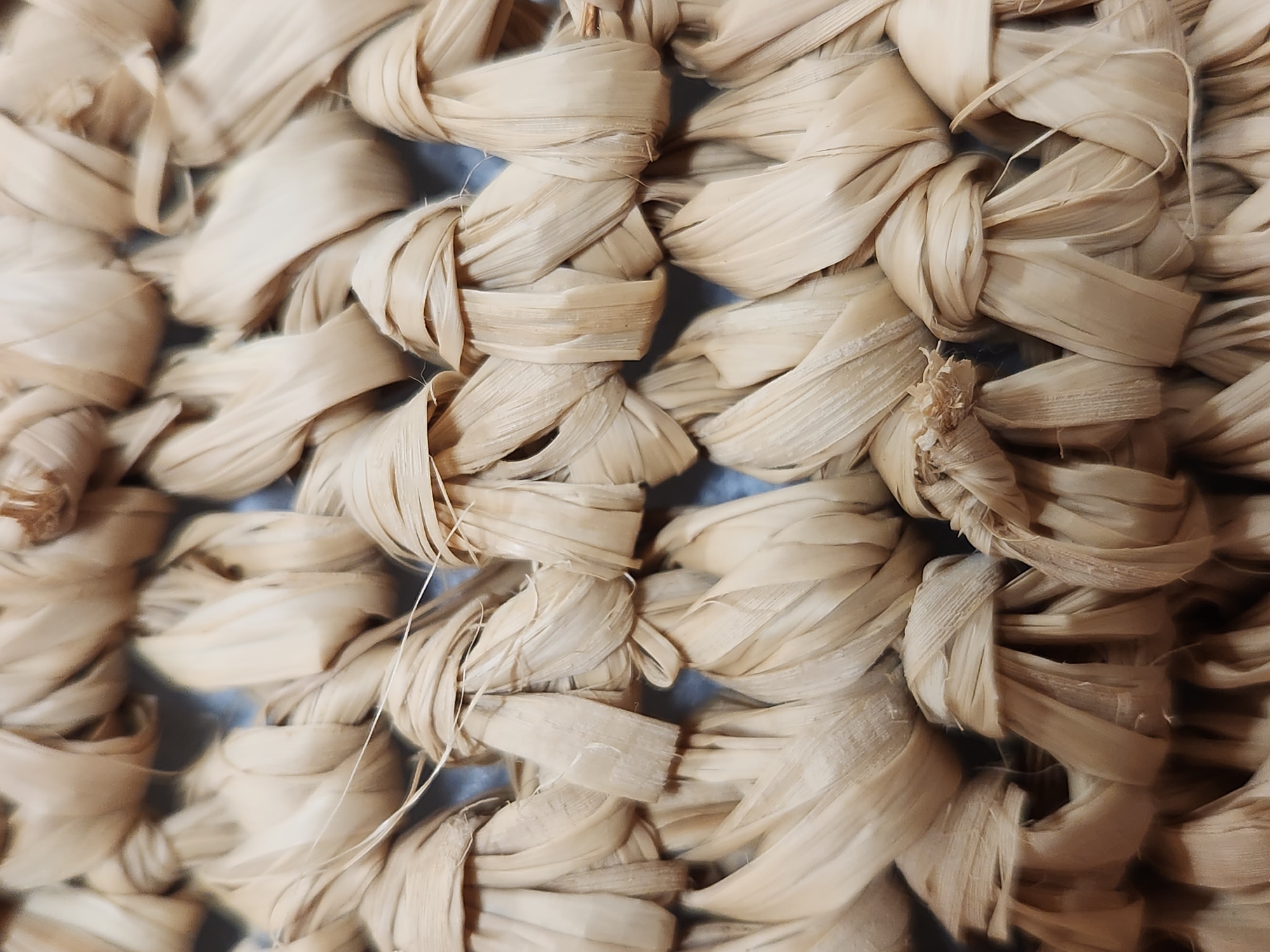 This image is a close-up, overhead shot of a woven straw-like material, possibly a basket or placemat. The texture is intricate, with strands of straw twisted and knotted together to form a repeating pattern. The color is a natural, light beige. The lighting is soft and even, highlighting the three-dimensional texture of the weave. There are no people, animals, or any other objects visible in the frame, nor is there any indication of a specific location within Maitland, United States of America. The focus is entirely on the detailed pattern and texture of the woven material.