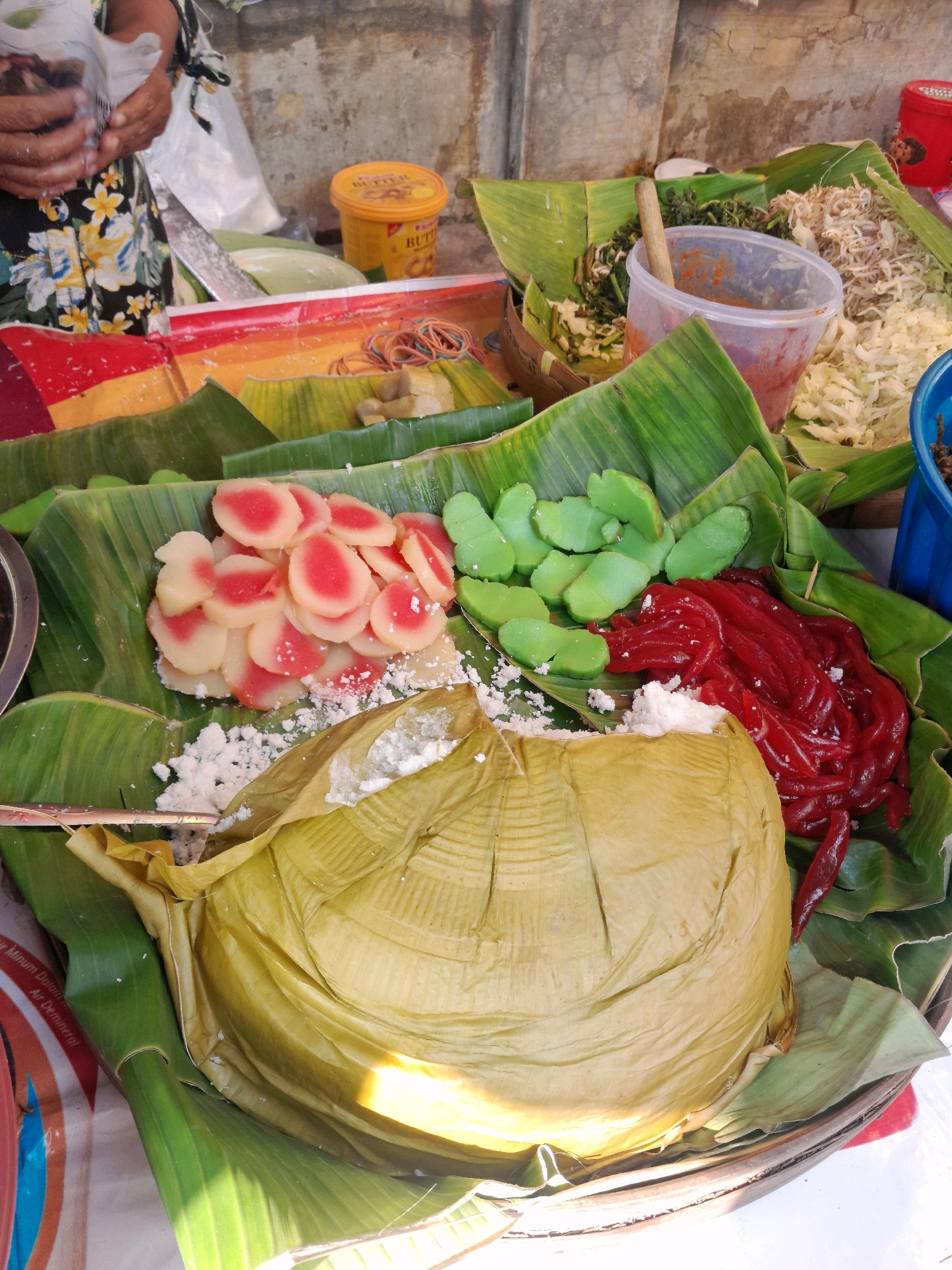 This image captures a street food stall in Jogosatru, Indonesia, focusing on a vibrant display of colorful desserts and accompaniments, likely prepared for sale. The foreground is dominated by a large, folded banana leaf parcel, appearing to contain a dish with a soft, fluffy interior, possibly steamed rice or a sweet cake, sprinkled with white shredded coconut. To the left of this parcel are slices of a pink and white food item, resembling candied fruits or sweet potato, also dusted with coconut. Adjacent to these are bright green, leaf-shaped sweets, and to the right, a mound of thin, red, jelly-like strands, possibly agar-agar or sweetened grass jelly. The food is artfully arranged on banana leaves, serving as both a natural platter and a decorative element. In the background, glimpses of other food items are visible, including shredded vegetables and what appears to be a spicy condiment in a plastic container, with a wooden spoon resting inside. A person's hands, wearing a floral patterned apron, are partially visible on the left, suggesting they are the vendor, possibly in the process of serving or arranging the food. The stall is set against a rustic, textured wall, common in outdoor Indonesian markets. The overall impression is one of a bustling, authentic local food scene, showcasing traditional Indonesian culinary delights.