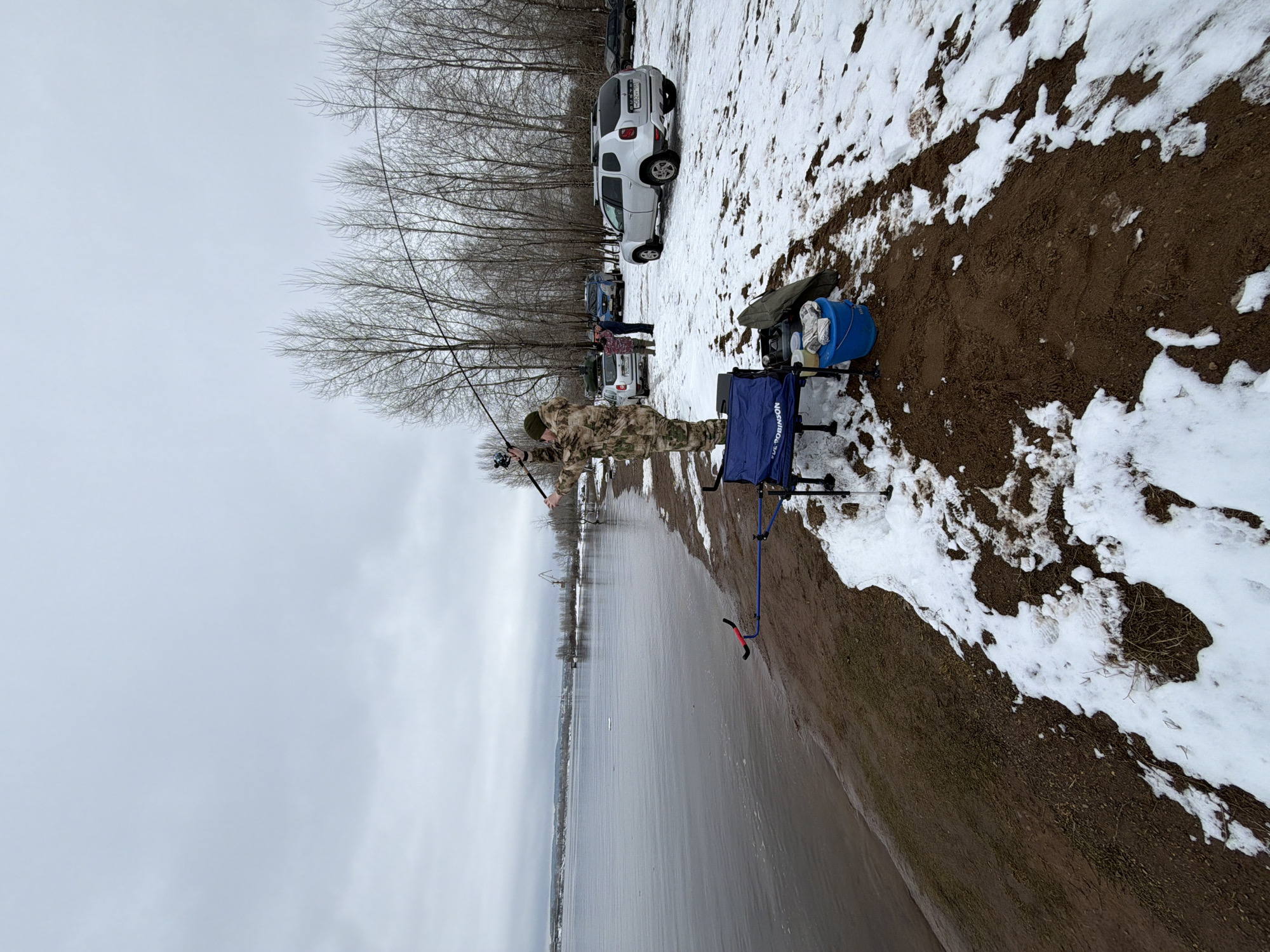 The image shows a person fishing on the shore of a body of water. The location is Chaikovsky, Russia. The person is dressed in camouflage clothing and holding a fishing rod. Next to them is a blue chair and other fishing equipment. The ground is a mix of dirt and snow. In the background, there are trees and several cars parked nearby. The sky is overcast. Several other people are standing near the vehicles.