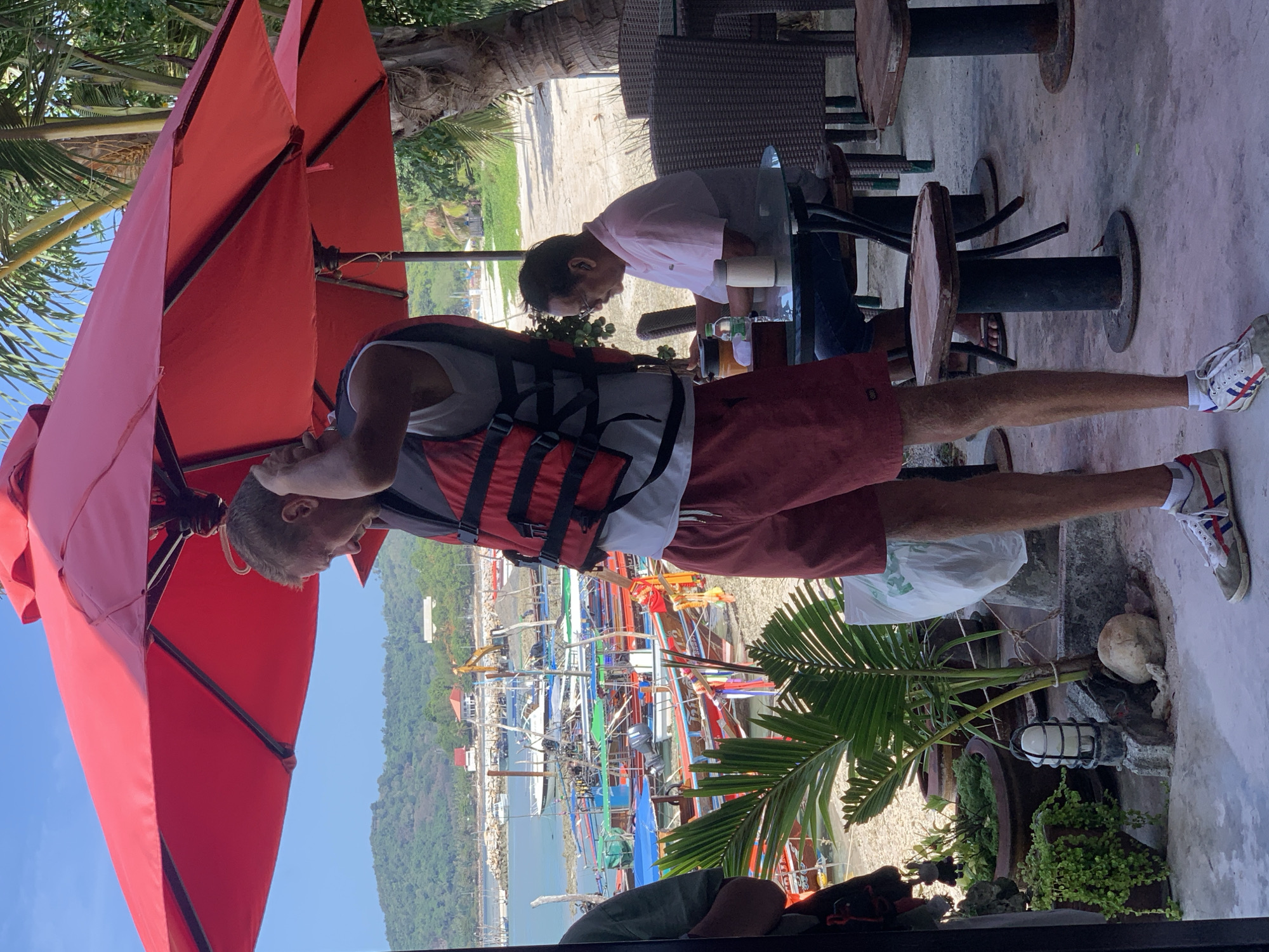 This image depicts an outdoor scene on a sunny day, likely at a beachside establishment in Ko Samui District, Thailand. In the foreground, a young man wearing a red and black life vest, white t-shirt, and reddish-brown shorts stands facing away from the camera, adjusting his hair. He is wearing white sneakers with red and blue accents. To his right, a mature man with glasses, wearing a pink and grey polo shirt and blue jeans, is seated at a round glass-top table. He is looking down at his hands. The table has a bottle of water and what appears to be a thermos. Behind the seated man, there are woven chairs and tables, suggesting a seating area for patrons.
The background reveals a view of a beach with numerous long-tail boats moored in the shallow water. Beyond the boats, there is a body of water leading to a coastline with green, hilly terrain and some buildings. The scene is partially shaded by a large red umbrella. Lush green plants, including a palm tree and potted plants, are visible in the lower left portion of the image, indicating a tropical environment. The overall impression is of a relaxed, possibly tourist-oriented location by the sea.
