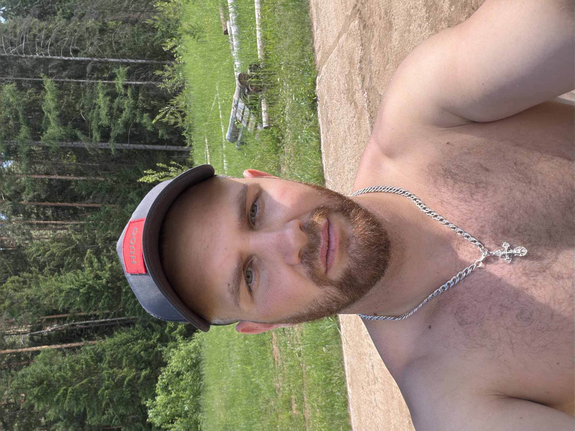 In the city of Prikamskiy, Russia, a man is pictured in a selfie taken outdoors during the day. He is wearing a grey baseball cap with a red "HUGO" logo. The man has light skin, green eyes, and a brown beard. He is shirtless and wearing a silver chain with a cross pendant. The background consists of a dense forest of pine trees and a grassy field. The ground in the foreground appears to be made of concrete or paving stones.