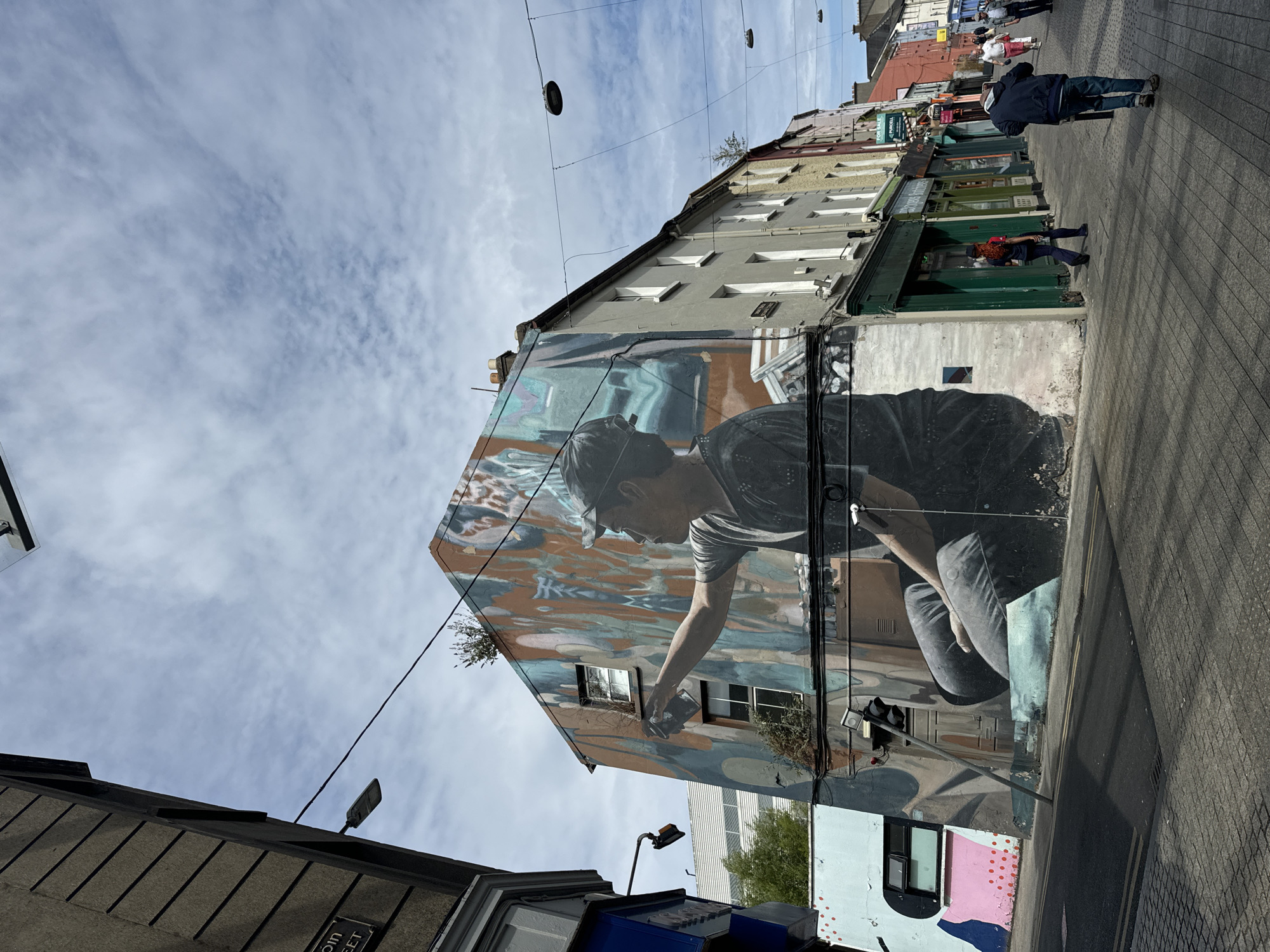 This is a wide-angle, eye-level shot of a street in Waterford, Ireland, featuring a large mural on the side of a building. The mural depicts a person wearing a cap and t-shirt, appearing to spray paint. The street is paved with cobblestones and asphalt, with a traffic light on the left side of the image. Several shops are visible along the right side of the street, with awnings and signs. A few people are walking along the sidewalk. The sky is cloudy, with patches of blue. The mural is highly detailed and colorful, with abstract elements surrounding the central figure. The building with the mural is a corner building, with its side facing the street. The overall impression is of a lively urban environment with artistic expression. The specific location is Reginald's Tower on the corner of Georges Street and Bailey's New Street in Waterford, Ireland.
