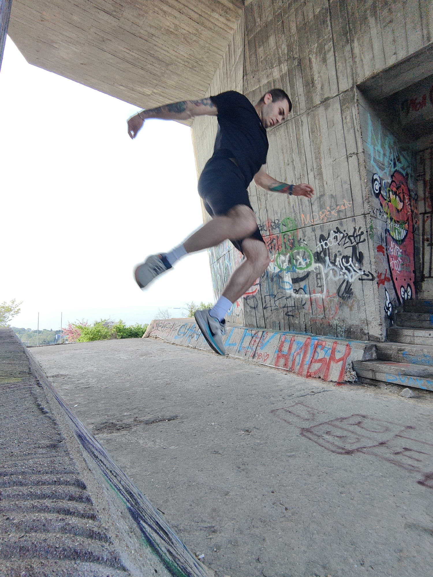 A man is captured mid-air, jumping, against a backdrop of an underpass with graffiti-covered walls. He is wearing a dark t-shirt, dark shorts, white socks, and light-colored running shoes. His left arm is extended, and his right arm is bent at the elbow, positioned slightly behind him. He appears to have tattoos on his left arm.
The location appears to be the underpass next to the "Monument of Bulgarian-Soviet Friendship" in Varna, Bulgaria. The underpass is constructed of concrete and is covered in graffiti. To the left of the man, there is a visible embankment with some greenery and hints of a distant cityscape or coastline. The lighting suggests it is a bright day.