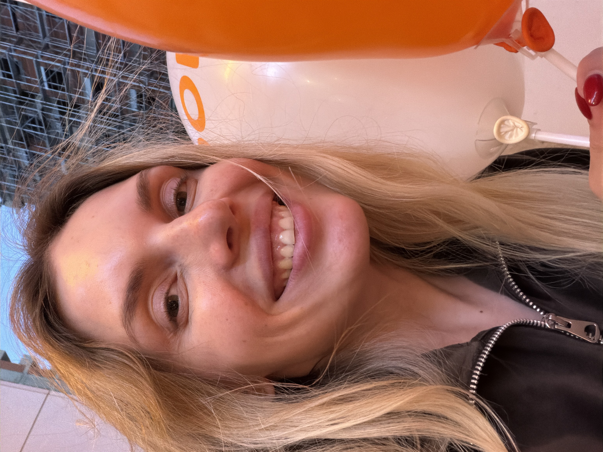 A young woman smiles at the camera while holding balloons, in what appears to be an urban setting. The balloons are orange and white. The woman has blonde hair and wears a dark-colored jacket with a visible zipper. The background shows architectural elements, possibly of a building. There is no definitive indication that the location is Budva, Montenegro, as the image lacks landmarks or features specific to that city.