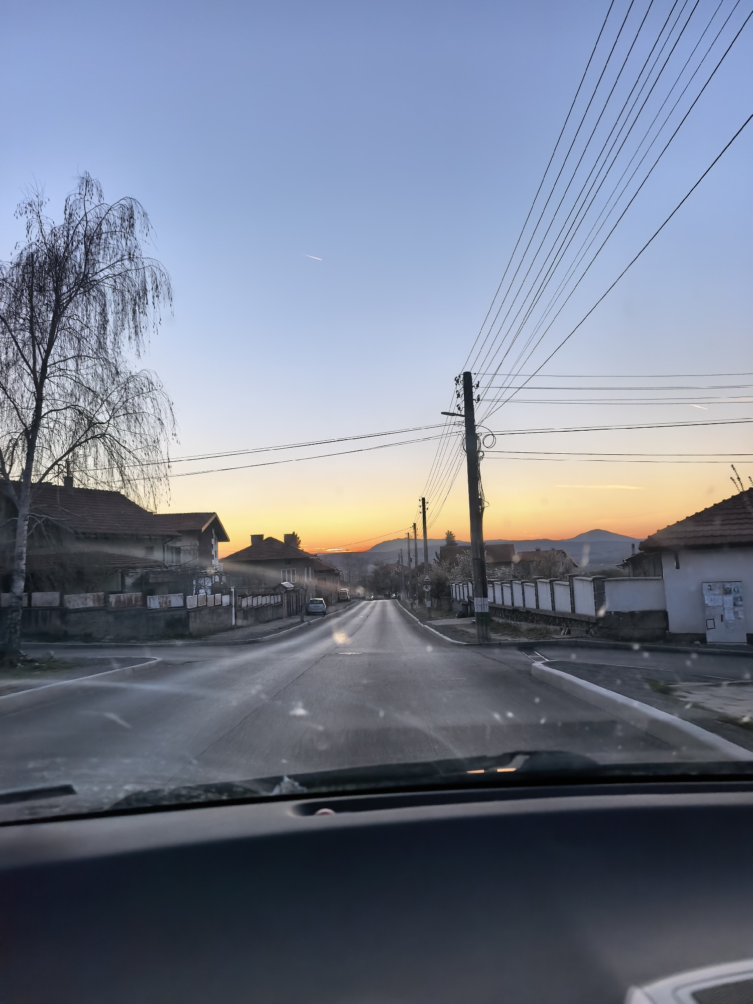 Captured from inside a vehicle, the image presents a street in a location designated as Unknown, Unknown, during what appears to be either sunrise or sunset. The street is paved and runs linearly into the distance, flanked on both sides by residential buildings. These buildings are a mix of single and two-story structures, many of which have perimeter walls or fences. Overhead, electrical wires stretch across the street. In the distance, faint hills can be seen on the horizon, contributing to the depth of the scene. To the left of the frame stands a bare tree. The foreground of the image, taken from within the car, shows the vehicle's dashboard and a partially obscured windshield.