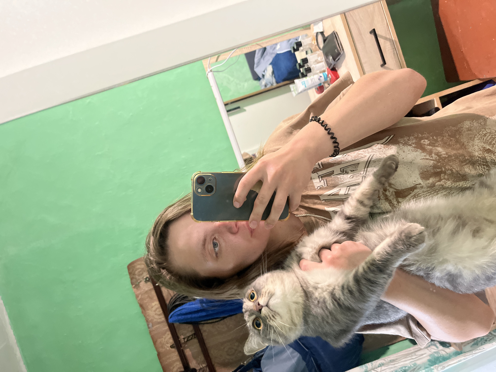 An image taken as a mirror selfie shows a young adult holding a cat in what appears to be an indoor setting in Nizhny Tagil, Russia. The perspective is from the mirror, showing the person holding a dark-colored smartphone, possibly an iPhone, to capture the reflection.
The main subject is a person with light-colored hair and blue eyes, wearing a light brown or tan t-shirt with a faded graphic design. They have a black coil-style bracelet on their left wrist. The person's expression is neutral. Cradled in their arms is a fluffy, grey and white cat with striking bright yellow-orange eyes. The cat is looking directly at the camera with a wide-eyed, surprised or alert expression, its whiskers prominent.
The background, largely reflected in the mirror, features a prominent bright green wall on the left. To the right, a wooden shelf or cabinet is visible, holding various bottles, containers, and other small items. Below the shelf, pieces of clothing, including blue fabric, are discernible. Part of a white ceiling runs along the top of the reflection, and a segment of a brown, patterned wooden trunk or furniture item is visible in the lower right background. No discernible text is visible on the phone screen, clothing, or background objects. The lighting suggests an indoor setting, likely during the day, though no direct light source is visible.