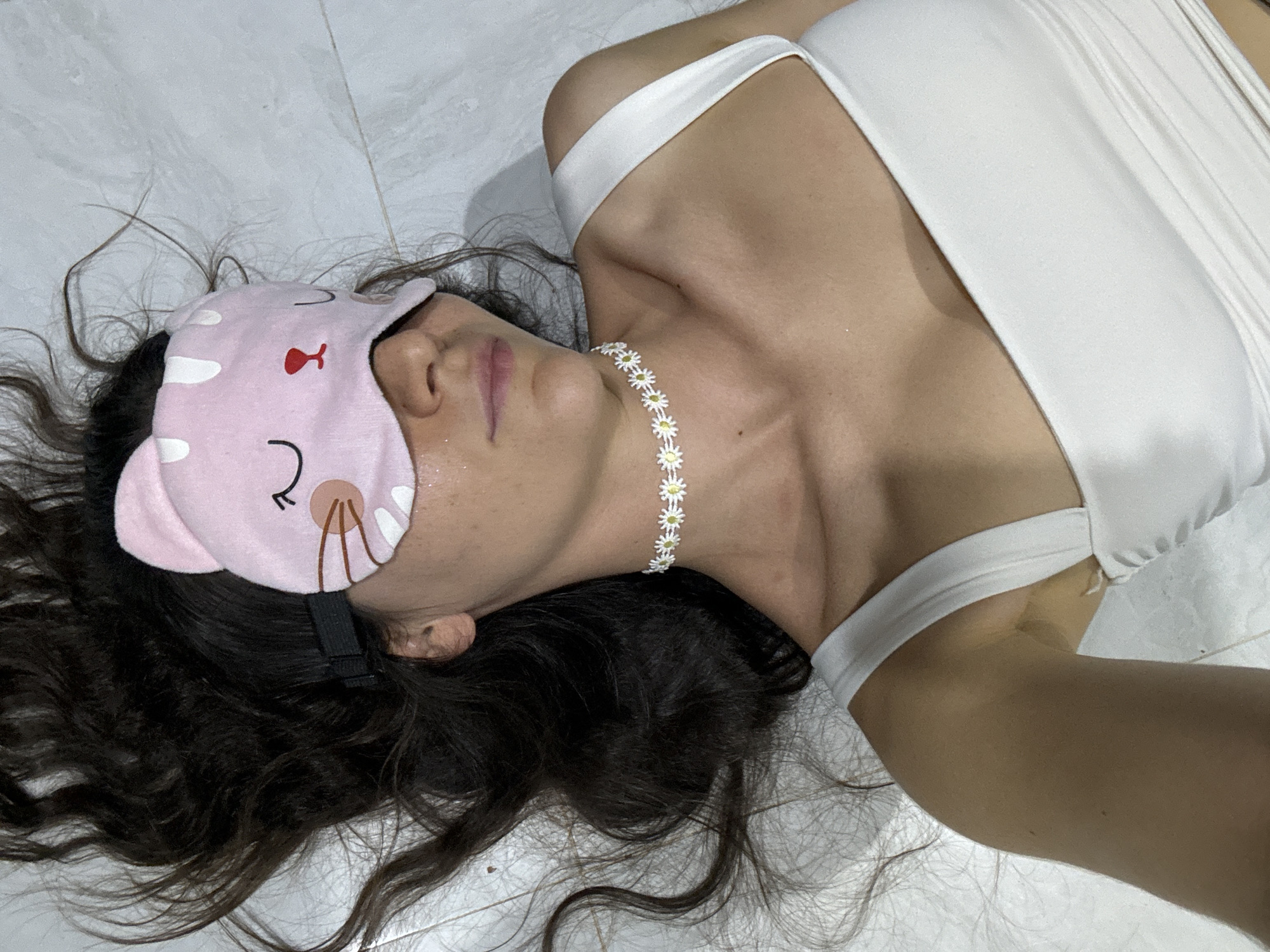 A person, likely a woman, is lying on a light-colored tiled floor, viewed from an overhead perspective, with her head oriented towards the upper left. She is wearing a pink, cat-themed sleep mask over her eyes, featuring white spots, drawn eyelashes, whiskers, and small ears. A white daisy-chain style choker necklace with yellow centers is visible around her neck. She is dressed in a white, possibly satin or silky, top or dress with a strap visible over her right shoulder. Her long, dark, wavy hair is spread out on the floor around her head.
The scene depicts an indoor setting, with the floor composed of light grey or white tiles with visible grout lines. The person's face beneath the mask appears relaxed, with her mouth slightly open. There is a subtle sheen on her skin, particularly around her nose and upper lip. The overall lighting is soft and even. No specific activity beyond lying down or resting is clearly indicated. No text is visible in the image. The location context provided for this image is Mũi Né, Vietnam.