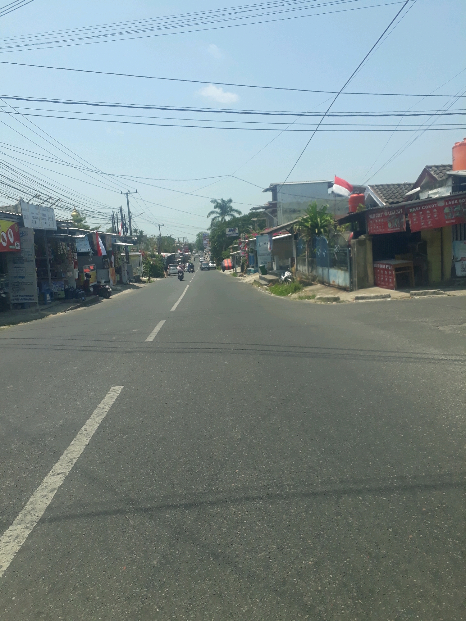The image captures a street scene in Pangkalpinang, Indonesia, likely on a sunny day given the clear blue sky. The road is asphalt with a white dashed line dividing the lanes. On the left side of the road, there are several small shops and stalls with awnings, displaying various goods. Some signs are visible, one with red lettering, and another appears to be an advertisement for "JNE". A person is sitting on a motorcycle in front of one of the shops. Further down the street, there are more buildings and more shops. On the right side of the road, there are also shops and stalls. One stall has red banners with white text, possibly advertising food. An Indonesian flag, red and white, is visible flying from a pole. Traffic, including a motorcycle and a few cars, is moving along the road in the distance. Overhead, a dense network of electrical wires crisscrosses the sky. A water tank is visible on top of one of the buildings. The general environment suggests a commercial or residential area with businesses lining the street.