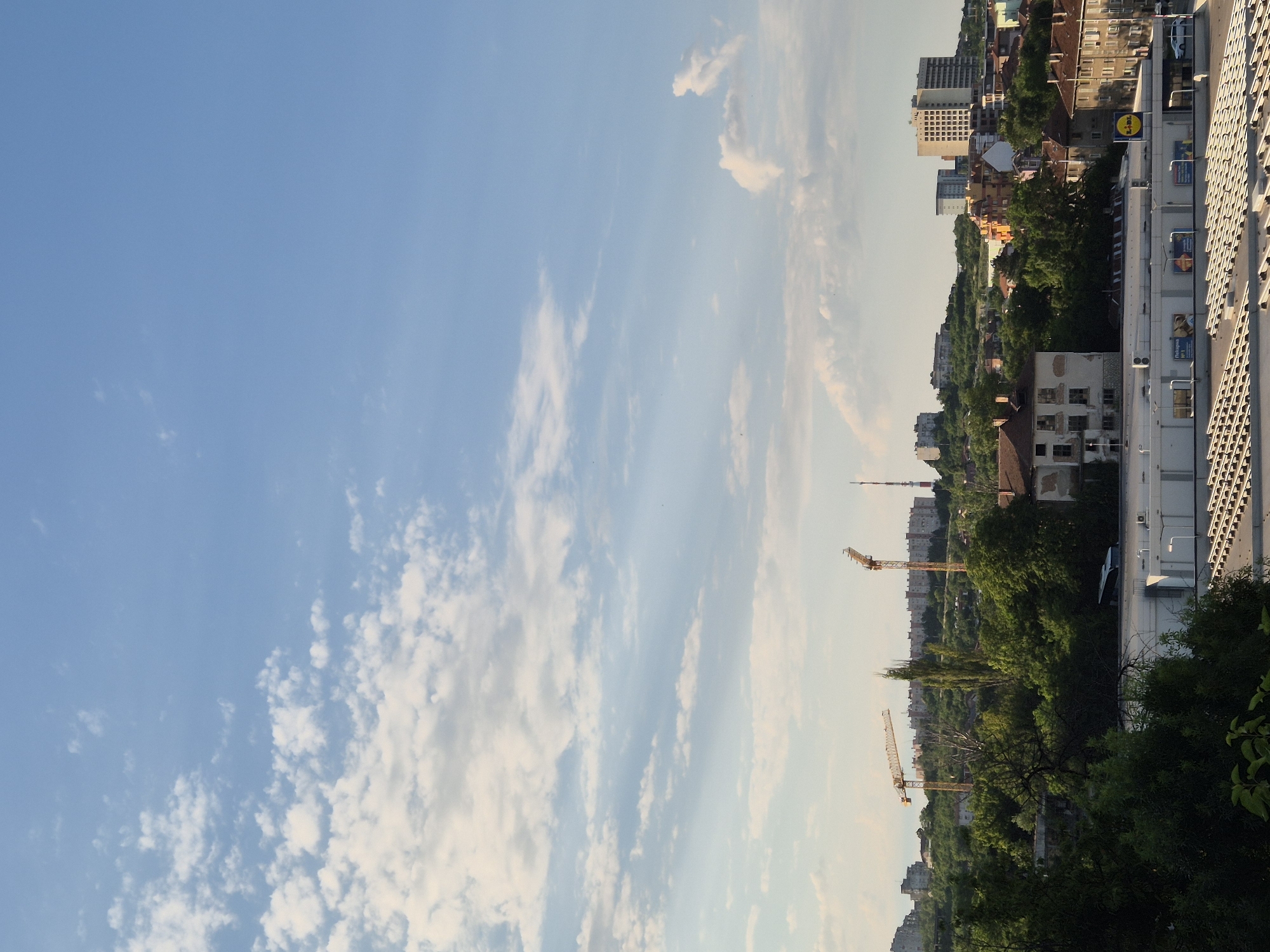 The image shows a cityscape under a bright blue sky with scattered white clouds. In the foreground, the rooftops of several buildings are visible, including a white building with a white awning and rows of rectangular structures. A supermarket with a yellow and blue LIDL logo is also present. Further in the background, there are trees and a cluster of buildings. Two yellow construction cranes are visible amidst the greenery and buildings. A bus is parked near one of the buildings. The scene appears to be an urban environment with a mix of residential and commercial structures, as well as ongoing construction.