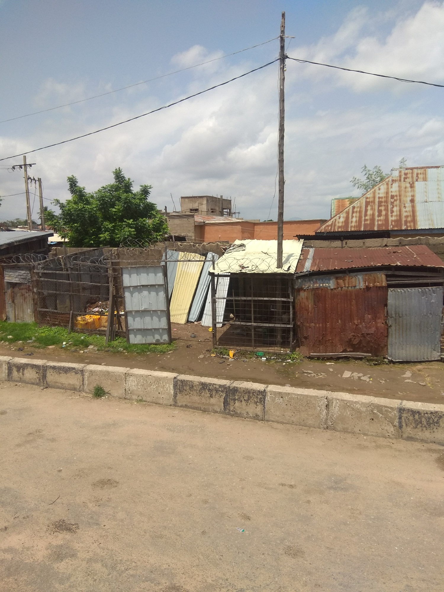 The image captures a street scene in Jalingo, Nigeria, focusing on a collection of informal structures. The foreground shows a paved road with a concrete curb. Beyond the curb, a patch of dry ground and sparse green vegetation leads to a fenced area. The fence is constructed with corrugated metal sheets, chain-link fencing, and topped with barbed wire. Inside the fenced area, there are several makeshift structures, predominantly made of corrugated metal sheets, some of which appear rusted. One structure has a white roof. There are also piles of debris and what looks like a yellow container visible within the enclosure. In the background, a wooden utility pole with wires extends upwards into a cloudy sky. Several buildings with corrugated metal roofs, some showing signs of weathering and rust, are visible behind the primary structures. A green tree stands to the left of the scene, and other buildings, some under construction, are further in the distance. The overall impression is of a developing urban environment with informal housing and commercial spaces.
