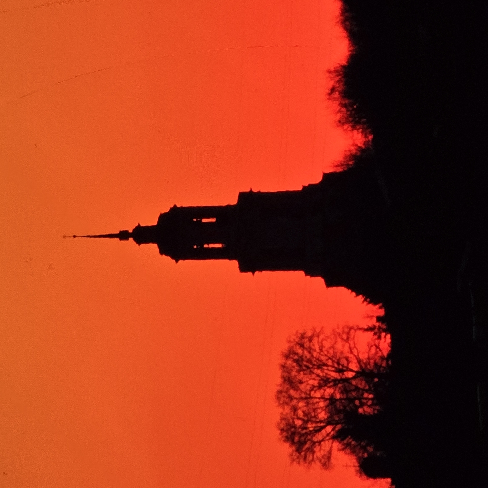 The image is a silhouette of a church steeple against a vibrant orange sky. The church appears to be St. Sophia's Cathedral in Vladimir, Russia, known for its distinctive onion dome. The sky is a uniform, intense orange, suggesting a sunset or sunrise. The foreground is dark, obscuring any details of the ground or surrounding landscape, but hints of trees can be seen on the left and right sides of the frame, also silhouetted against the bright sky. There are no people or animals visible in the image. The overall impression is one of dramatic beauty, with the stark silhouette of the church creating a strong focal point.