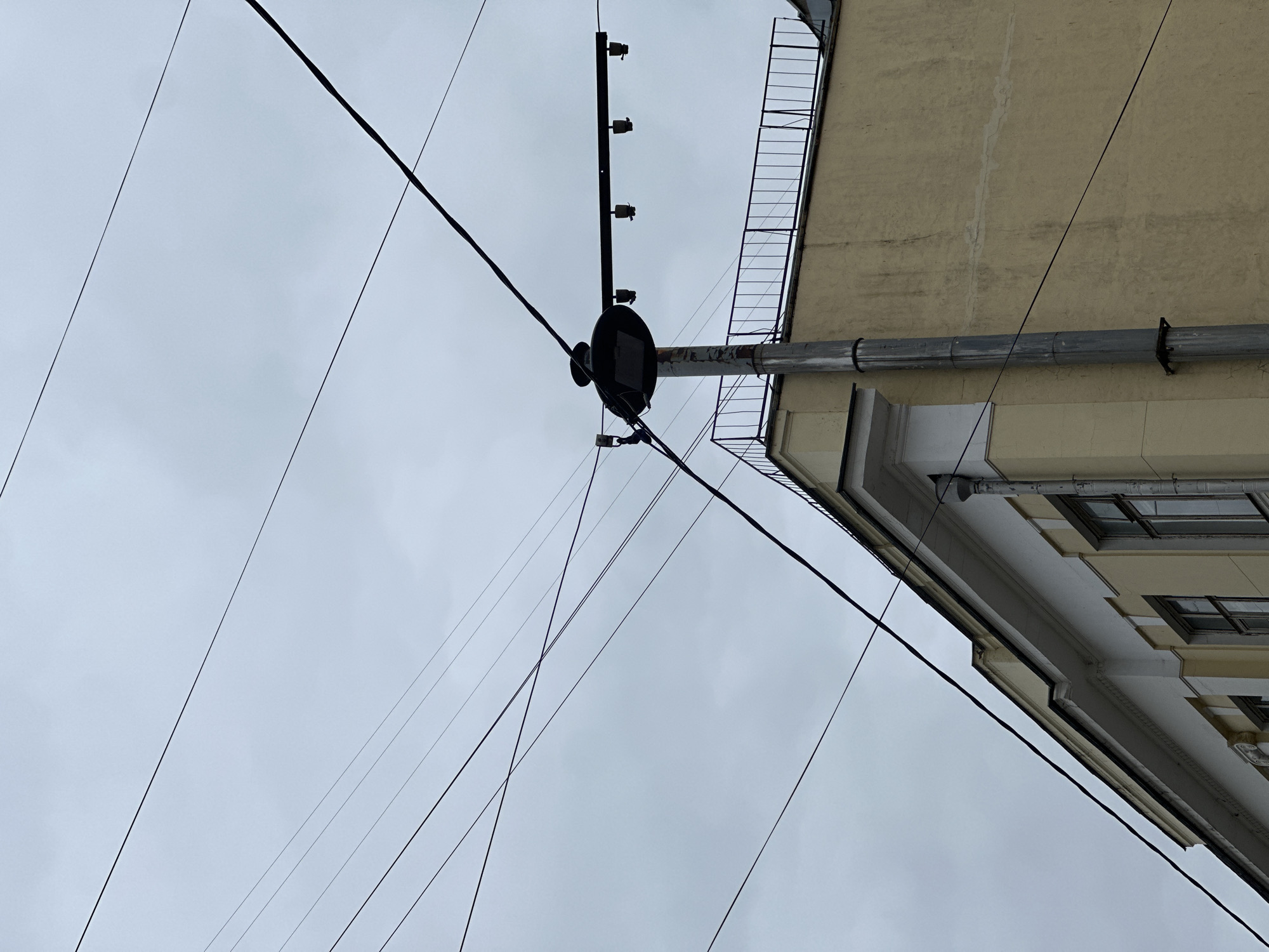 The image shows an upward-angle view in Moscow, Russia, depicting a building facade and electrical infrastructure against an overcast sky. The facade is of a light-colored building with architectural details including a white cornice, rectangular windows, and a grey drainpipe. A metal fire escape ladder is visible attached to the building. In the foreground, there is a utility pole with a circular junction box and a series of ceramic insulators on a vertical support. Multiple black electrical wires stretch across the sky, intersecting near the junction box. The sky is a uniform grey.