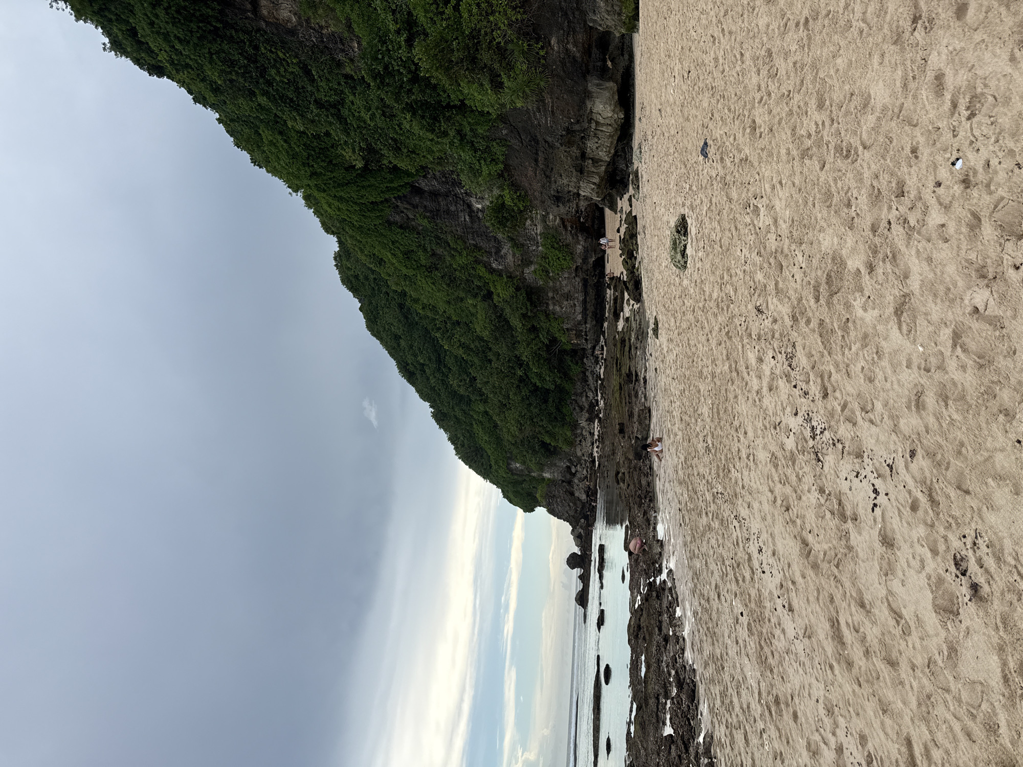 The image depicts a coastal scene in Ungasan, Indonesia. A rocky, vegetation-covered cliff dominates the left side of the image. A sandy beach stretches along the right, meeting the ocean at the bottom. Along the shoreline are dark rocks and visible water. The sky is overcast, with varying shades of gray. Two people are visible on the beach: one standing near the base of the cliff and the other bent over the water along the shoreline. The photo is a wide, landscape shot.