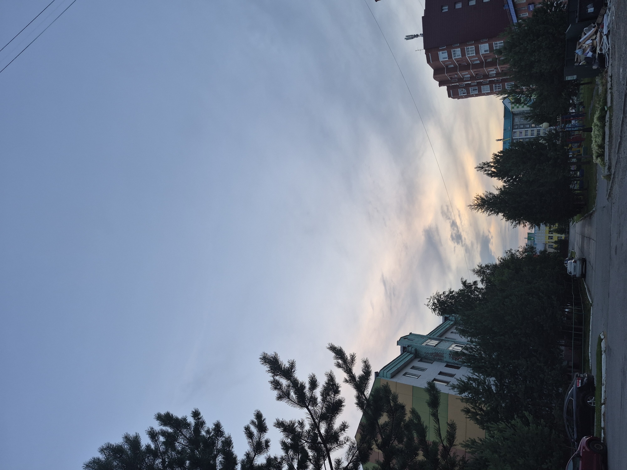 This is a vertical, eye-level, outdoor, daytime, full shot of a residential street in the city of Nyagan, Russia. The sky is a pale blue with streaks of clouds. In the foreground, on the left side of the frame, is the branch of a pine tree. To the right of the tree is a building with a colorful facade of green and yellow, with a turquoise roofline and several windows. Further down the street, on the left, are a row of dark green coniferous trees. Behind these trees are more buildings, including a light blue and white building with numerous windows. On the right side of the frame is a tall, reddish-brown apartment building with many windows and a cell tower on its roof. In front of this building is a patch of grass with a playground in the background. There are several cars parked along the street, including a red car on the left and a black sedan and a silver sedan further down. The street itself is paved with asphalt. There are power lines visible in the upper right portion of the image. The scene appears to be a quiet residential area in the late afternoon or early evening.