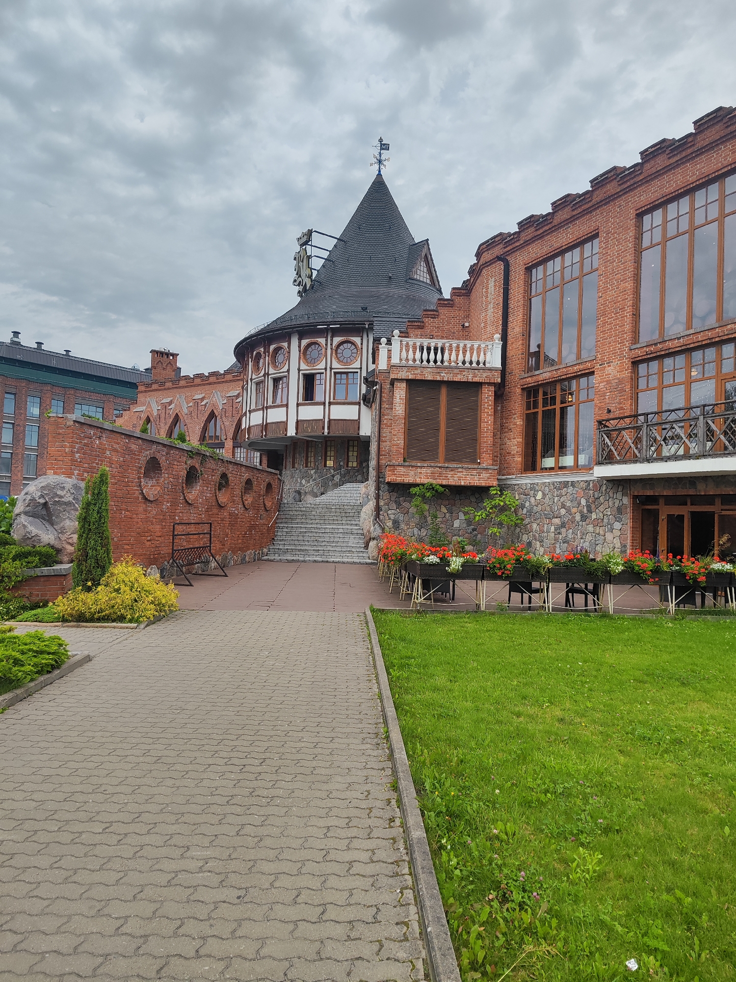 The image depicts the exterior of a building with a unique architectural style, resembling a castle or an old fort, situated in Kaliningrad, Russia. The building is constructed primarily of red brick, with a prominent conical dark roof and a circular section featuring arched windows. Adjacent to this structure is a taller, more modern brick building with large windows. In the foreground, a paved walkway leads towards the building. To the right of the walkway is a well-maintained green lawn with several outdoor tables and chairs set up in front of a stone-faced section of the building, adorned with planters filled with red and white flowers. To the left of the walkway, a brick wall with circular openings runs along a garden area that includes a large rock, a small evergreen tree, and yellow bushes. The sky is overcast with gray clouds. There are no people visible in the scene.