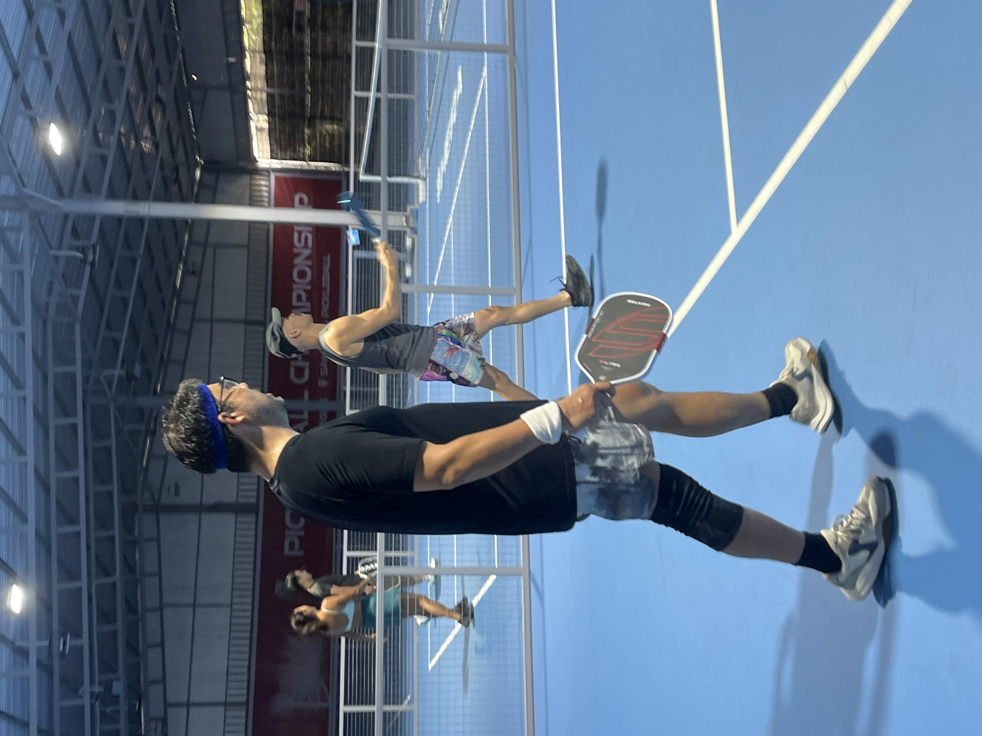 The image depicts an indoor pickleball court in Da Nang, Vietnam. The court is bright blue with white lines. In the foreground, a man wearing a black t-shirt, patterned shorts, a blue headband, glasses, and a knee brace is walking. He is holding a pickleball paddle. Behind him, another man in a grey tank top and colorful shorts is also holding a paddle and appears to be in motion. In the background, two women are visible on the court, one in a teal top and shorts, and the other in a dark shirt. The facility has a metal roof structure and a banner in the background with the text "PICKALL CHIP". The lighting appears to be artificial.