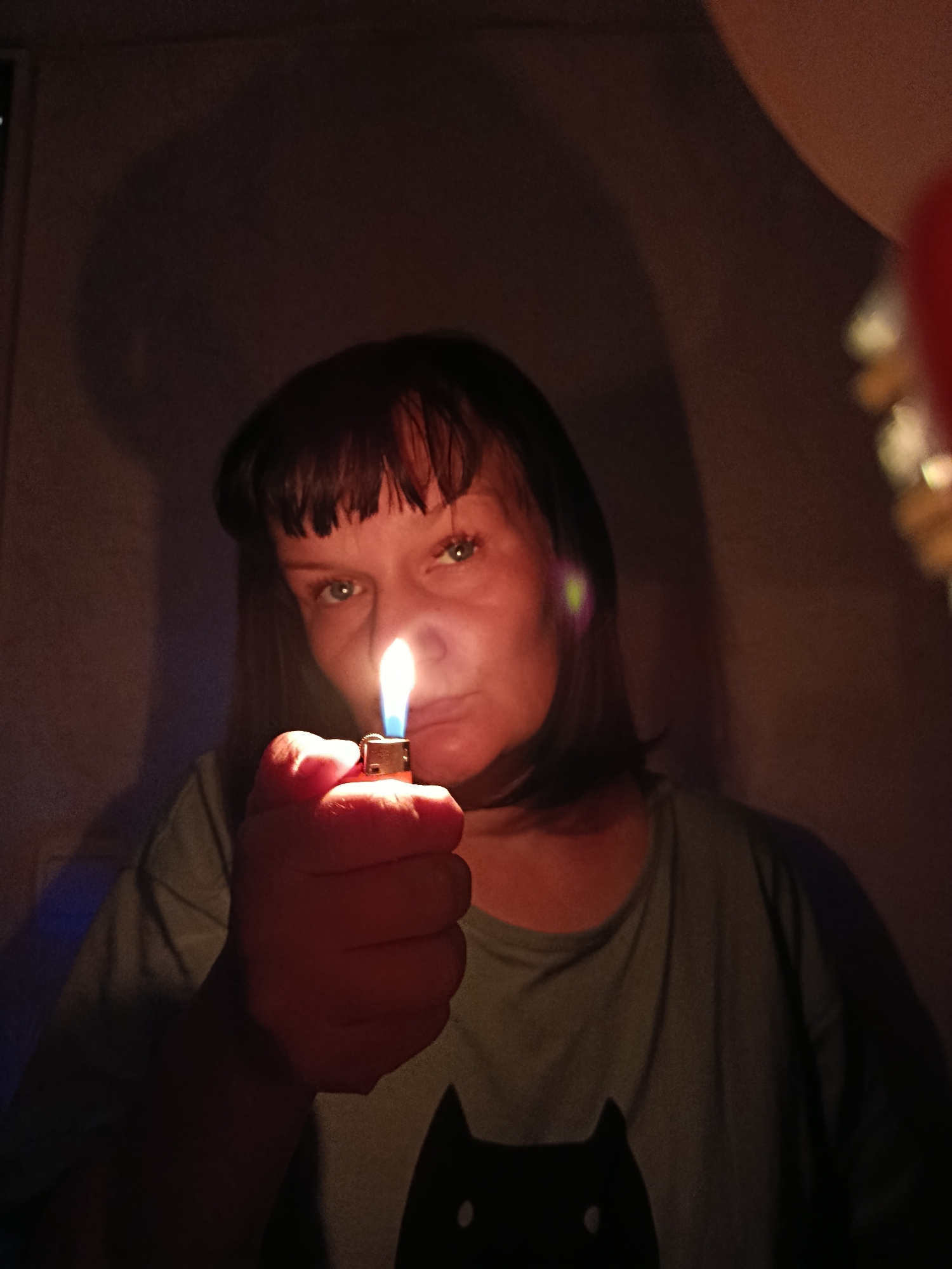 A woman is holding a lit lighter in front of her face in a dark room. She has short, dark hair and is wearing a t-shirt with a black cat design. The flame is illuminating her face and creating a shadow behind her. The location is unknown.