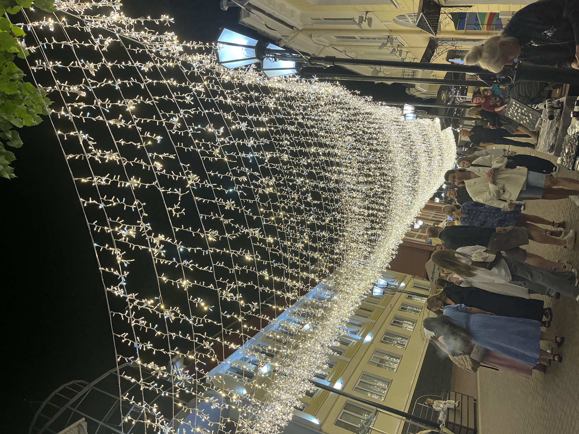 The image depicts a street scene at night, likely in Vitsebsk, Belarus, characterized by an abundance of twinkling fairy lights strung across the street, creating a canopy effect. The street is lined with buildings, one of which is illuminated with yellow light, featuring multiple windows. Several people are present, some walking and others standing in groups. Their attire suggests casual wear. In the foreground, a stall displaying various items, possibly jewelry or souvenirs, is visible, with a woman attending to it. Street lamps are also present, contributing to the illumination of the area. The overall atmosphere is festive and illuminated, suggesting a public space or thoroughfare, possibly a pedestrian zone.