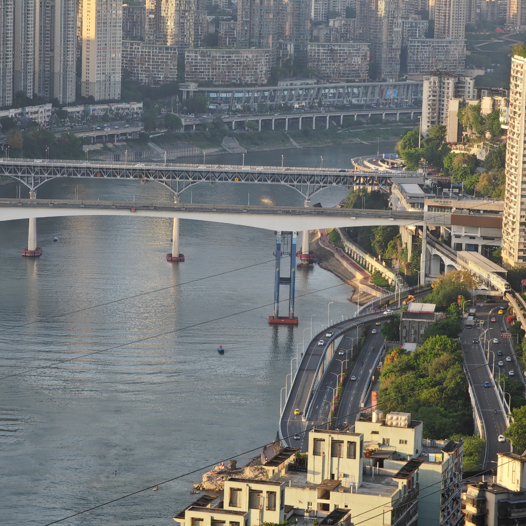 The image depicts a daytime aerial view of Yuzhong District in Chongqing, China, showcasing a river running through a dense urban environment. Multiple bridges span the river, with cars traveling across them. The river surface is relatively calm, and there are a few small boats. Buildings of varying heights line the riverbanks, creating a skyline of residential and commercial structures. Roads run alongside the river, with vehicles moving on them. Green vegetation is visible in some areas, juxtaposed against the concrete of the urban landscape. There are people gathering on the river’s edge. Electrical wires are visible in the foreground.
