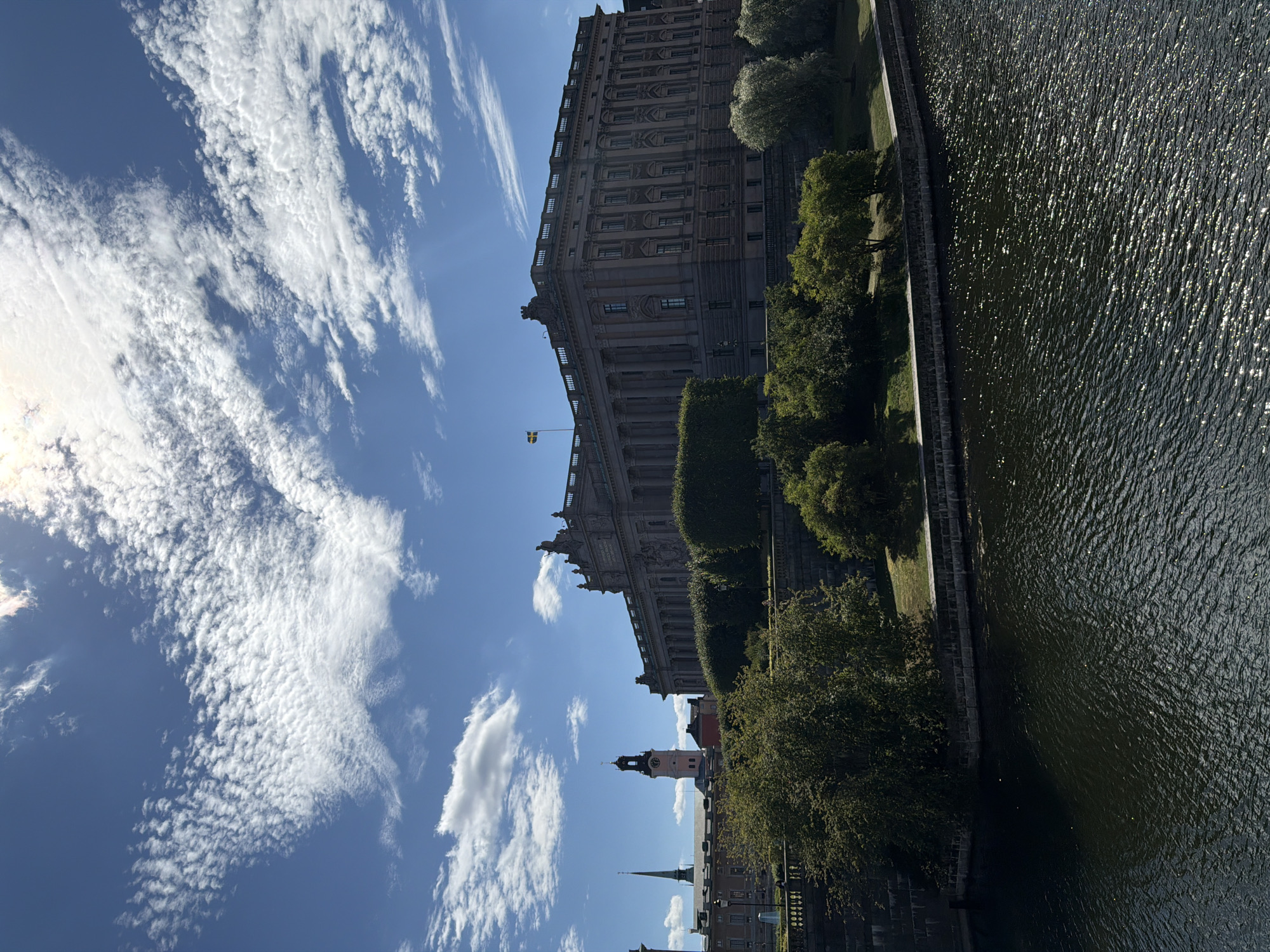 The image shows a wide, dark body of water in the foreground, with ripples and reflections of sunlight on its surface. On the right bank of the water, there is a stone embankment topped with manicured green trees and bushes. Behind the trees, a large, imposing stone building with classical architectural features, including a colonnade and decorative sculptures, dominates the middle ground. A Swedish flag flies from a flagpole on the roof of this building. To the left of the main building, there are other buildings visible, including a structure with a tall spire and a building with a clock tower that has a pink base and a dark clock face. The sky above is a vibrant blue, dotted with scattered white clouds, some of which appear feathery and wispy. The overall scene appears to be a bright, sunny day. This is a view of the Stockholm City Hall (Stadshuset), located on the island of Kungsholmen in Stockholm, Sweden, overlooking the waters of Lake Mälaren.