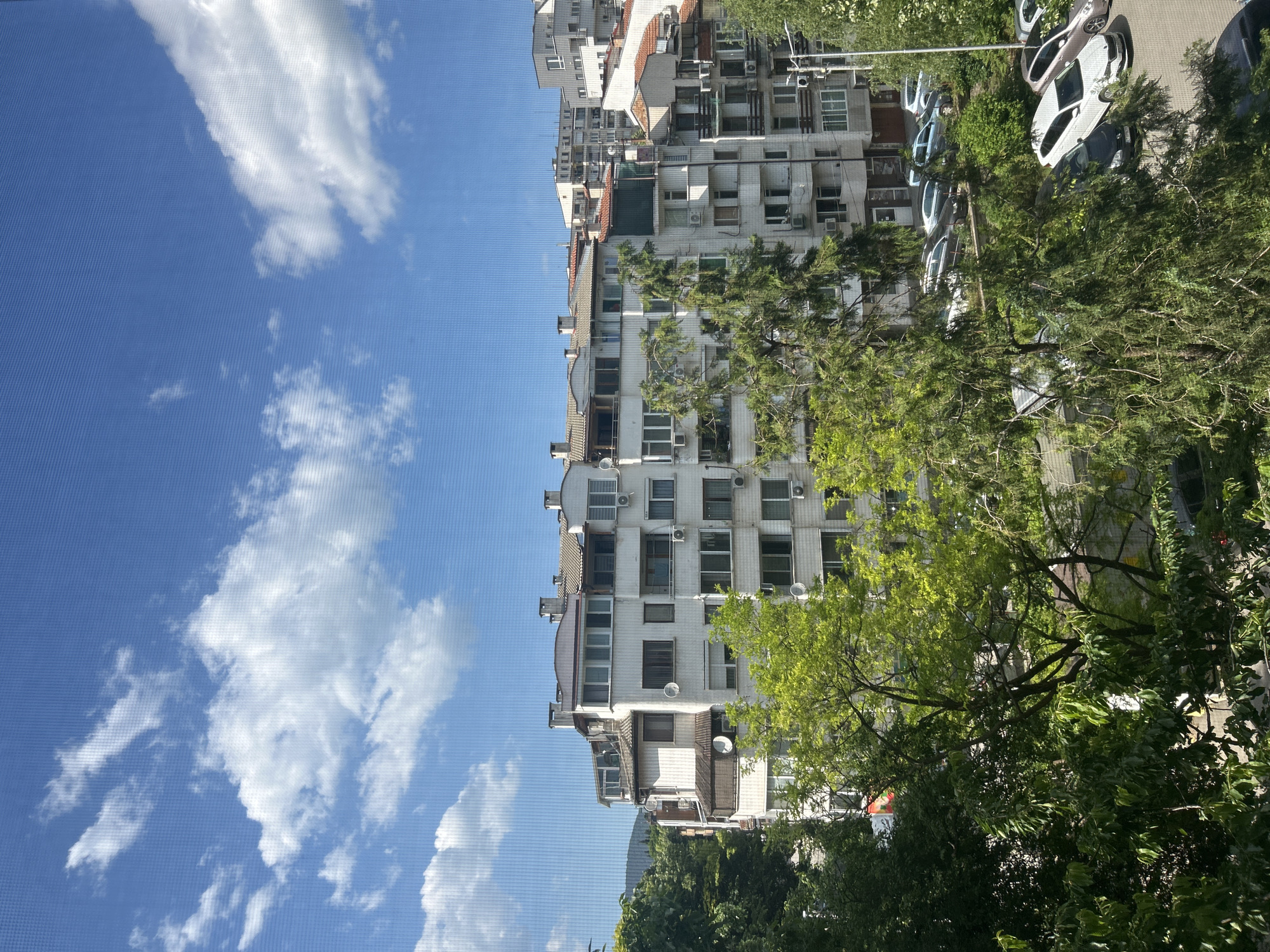 The image shows an apartment building under a partly cloudy sky. The building is densely packed with windows and air conditioning units. In front of the building are trees and parked cars. The location is likely an urban area in Unknown, Unknown.