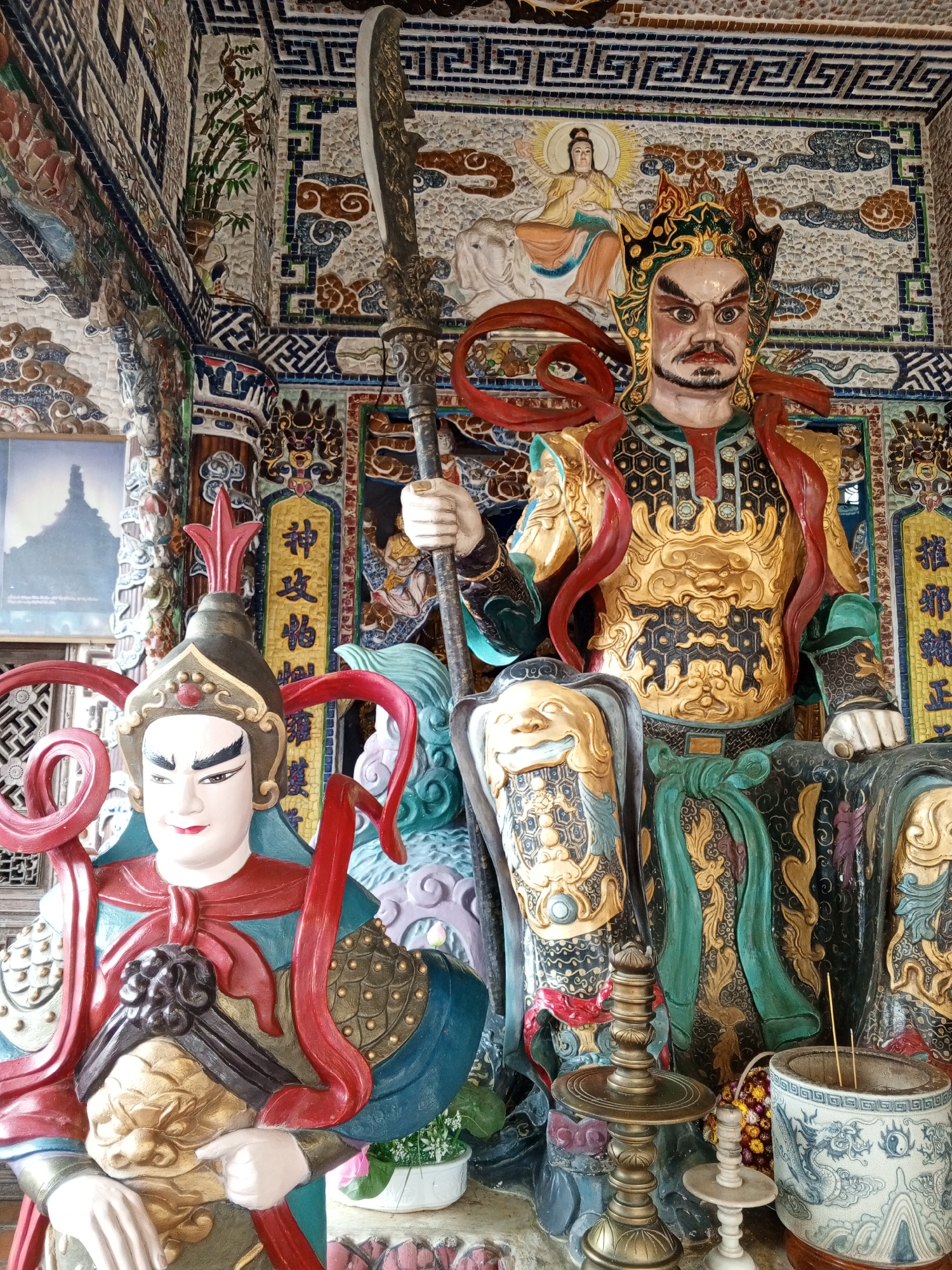 This is a medium shot, eye-level, outdoor photograph of religious statues within a temple in Xuan Thanh, Vietnam. The central subject is a large, elaborately decorated statue of a warrior, possibly a deity or guardian, with a stern expression, a golden breastplate adorned with intricate patterns and a dragon motif, and a red headdress with flowing ribbons. To the left, a second, smaller statue of a warrior with a similar headdress and costume is visible. Both statues are rendered in vibrant colors, including gold, red, blue, and green. The background is a richly decorated wall, also covered in intricate mosaic tiles depicting various motifs, including clouds, figures, and geometric patterns. A tall, ornate spear or pole is held by the larger statue. In the foreground, there are several objects, including a brass candlestick, a small white candlestick, and a decorative ceramic pot filled with incense sticks. A framed image or print of a building is visible on the left side of the image. The lighting suggests it is daytime.